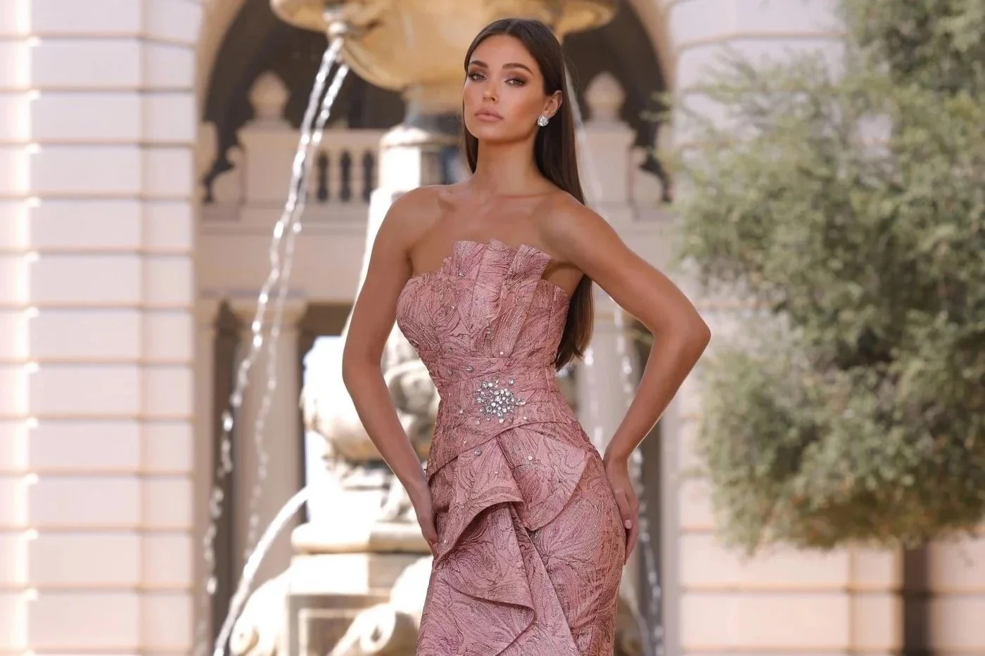 A woman wearing an elegant off-the-shoulder metallic gold and blue gown, standing in a wooden-paneled room with a window in the background.