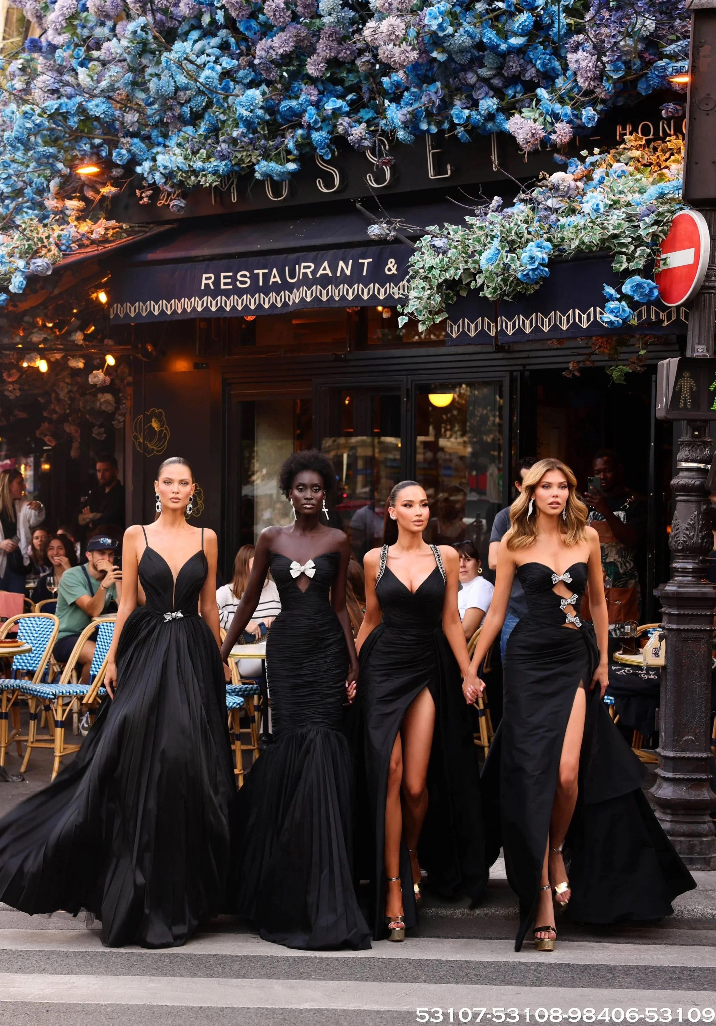 Four women dressed in elegant black evening gowns walking on a city street in front of a restaurant with outdoor seating. The women are holding hands, and onlookers are seated at tables behind them.