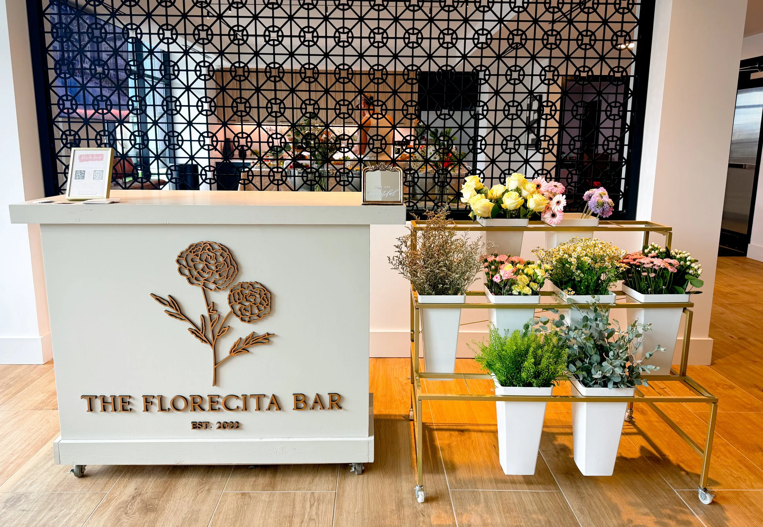 A flower shop reception with a white counter displaying a framed sign and floral arrangements on wheels. Behind the counter is a decorative black lattice screen, with flowers and plants visible in the background.