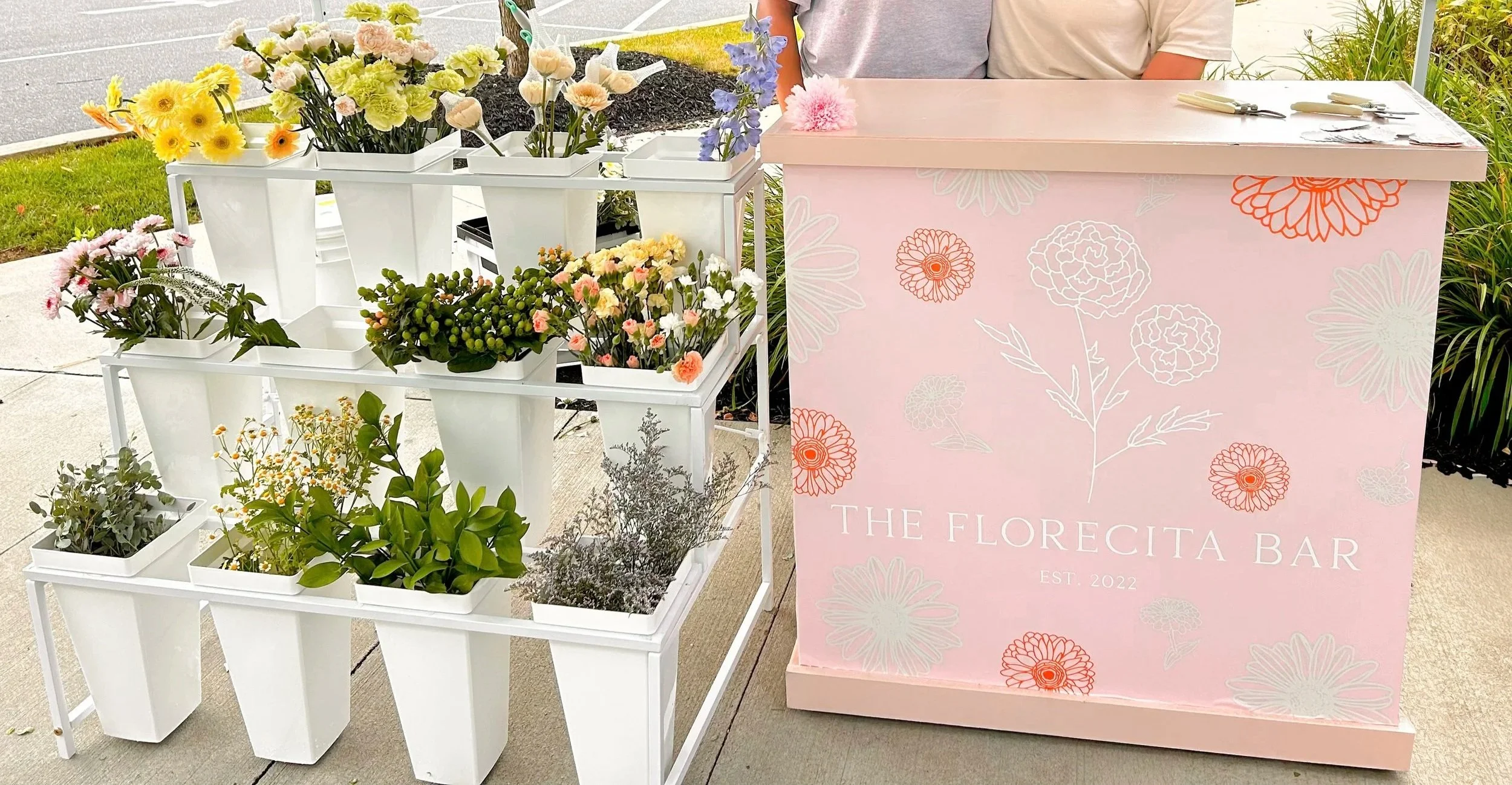A flower stand displaying various potted and cut flowers at an outdoor event, with a pink floral-themed sign reading "The Florecita Bar."