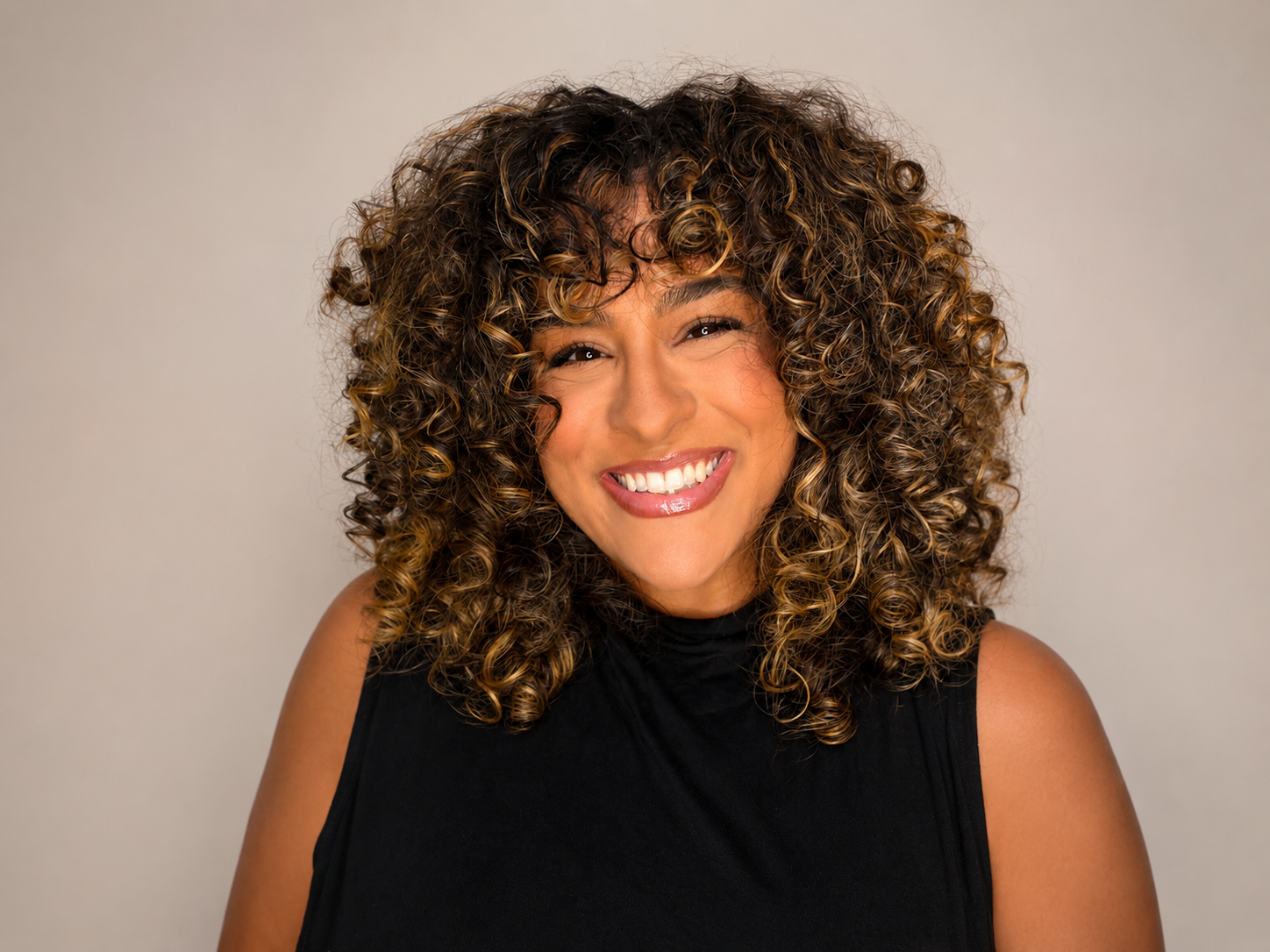 A woman with curly, voluminous hair smiling at the camera, wearing a sleeveless black top, against a plain light-colored background.