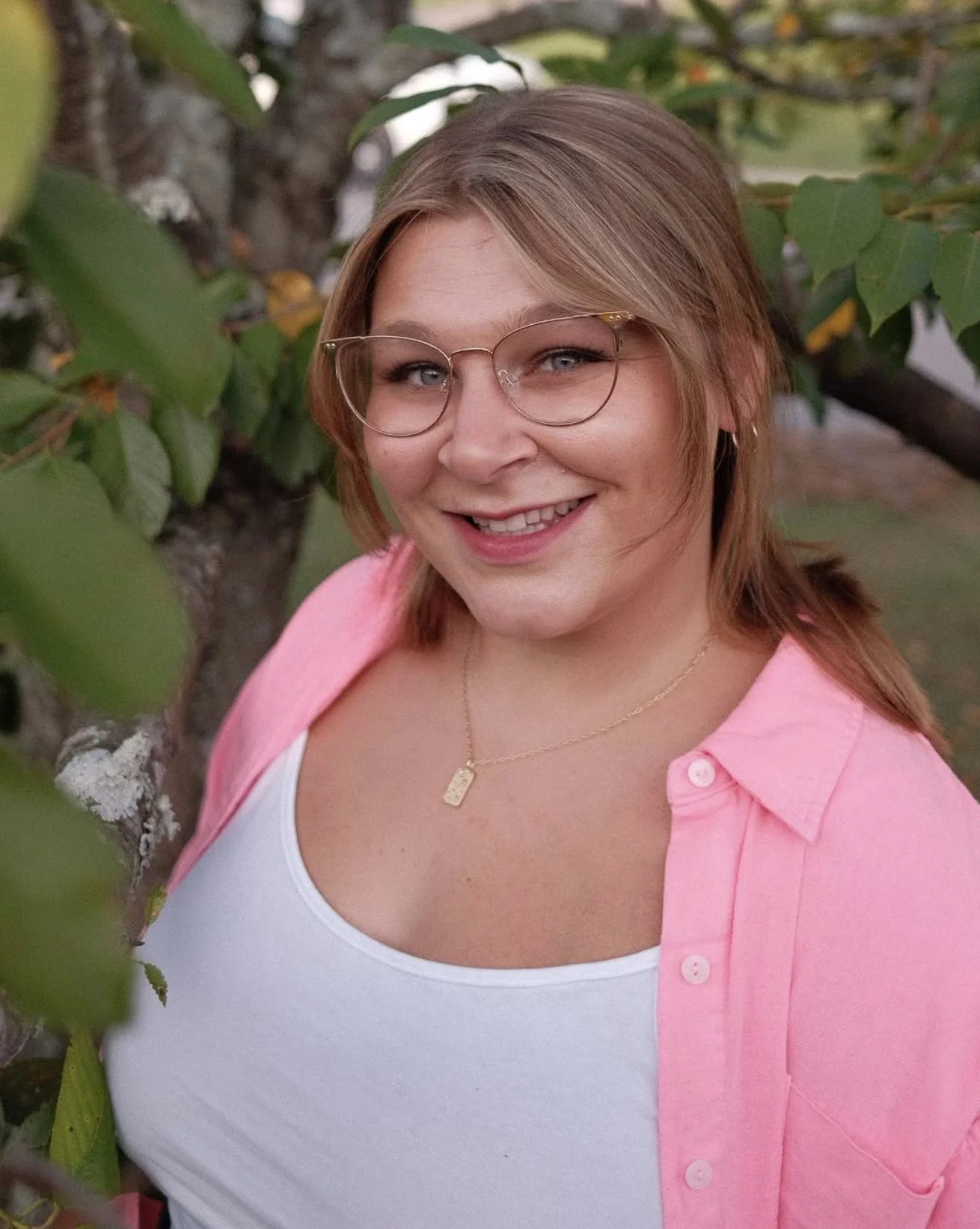 A woman with blonde hair, glasses, and a pink shirt smiling outdoors near a tree with green leaves