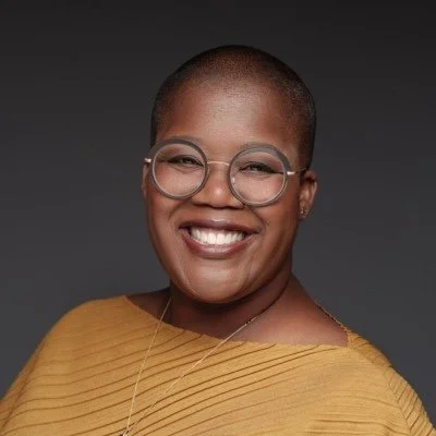 Portrait of a smiling Black woman with glasses and short hair, wearing a yellow top.