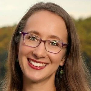 A smiling woman with long brown hair, glasses, and green earrings outdoors.