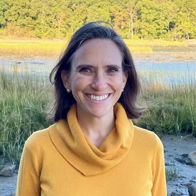 A smiling woman with shoulder-length brown hair wearing a yellow sweater outdoors near a body of water with trees in the background.