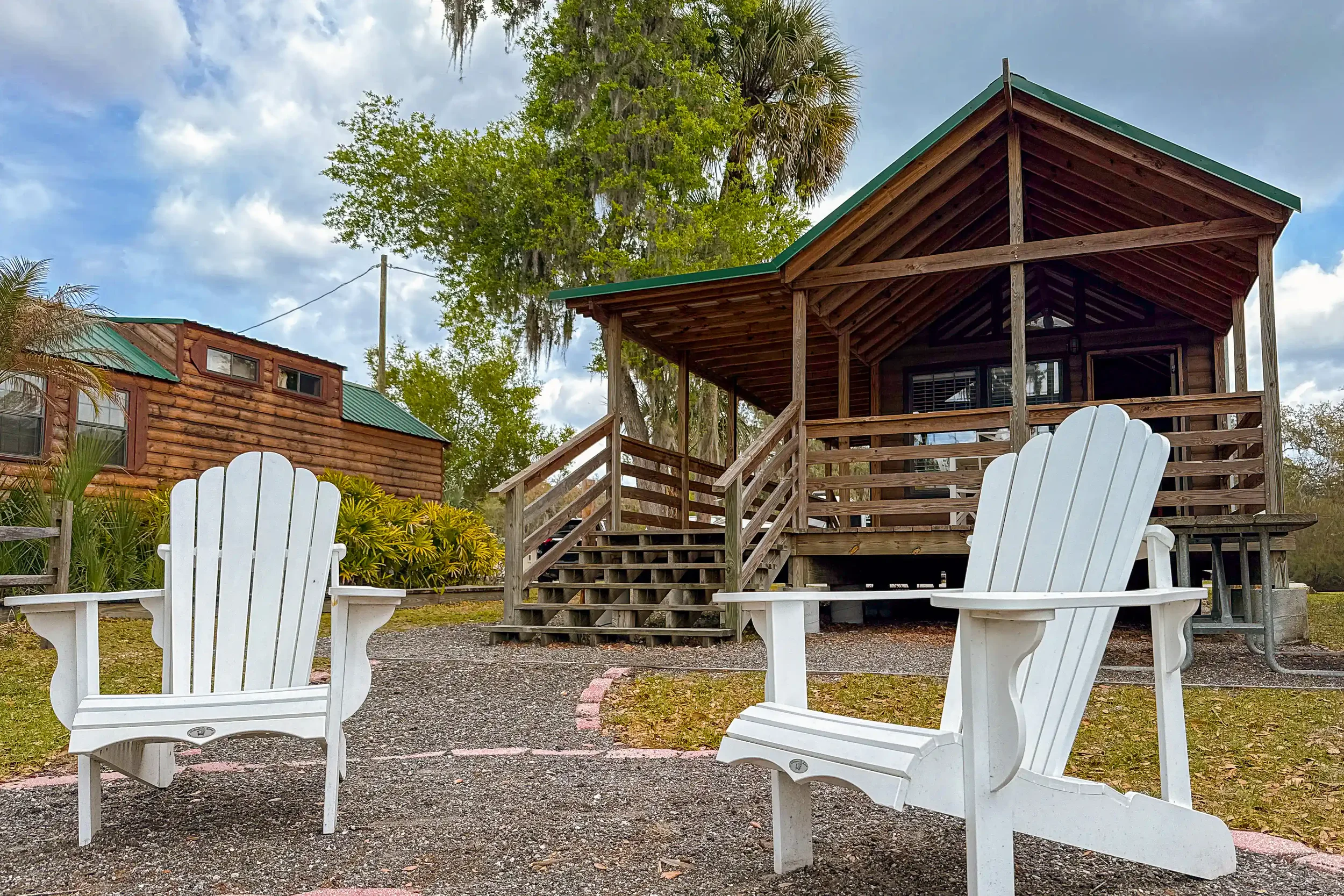 Two white Adirondack chairs sit in the foreground facing a rustic, wooden cabin with a green roof and a covered front porch. The cozy campsite is surrounded by large Old Florida trees draped in Spanish moss under a partly cloudy sky.
