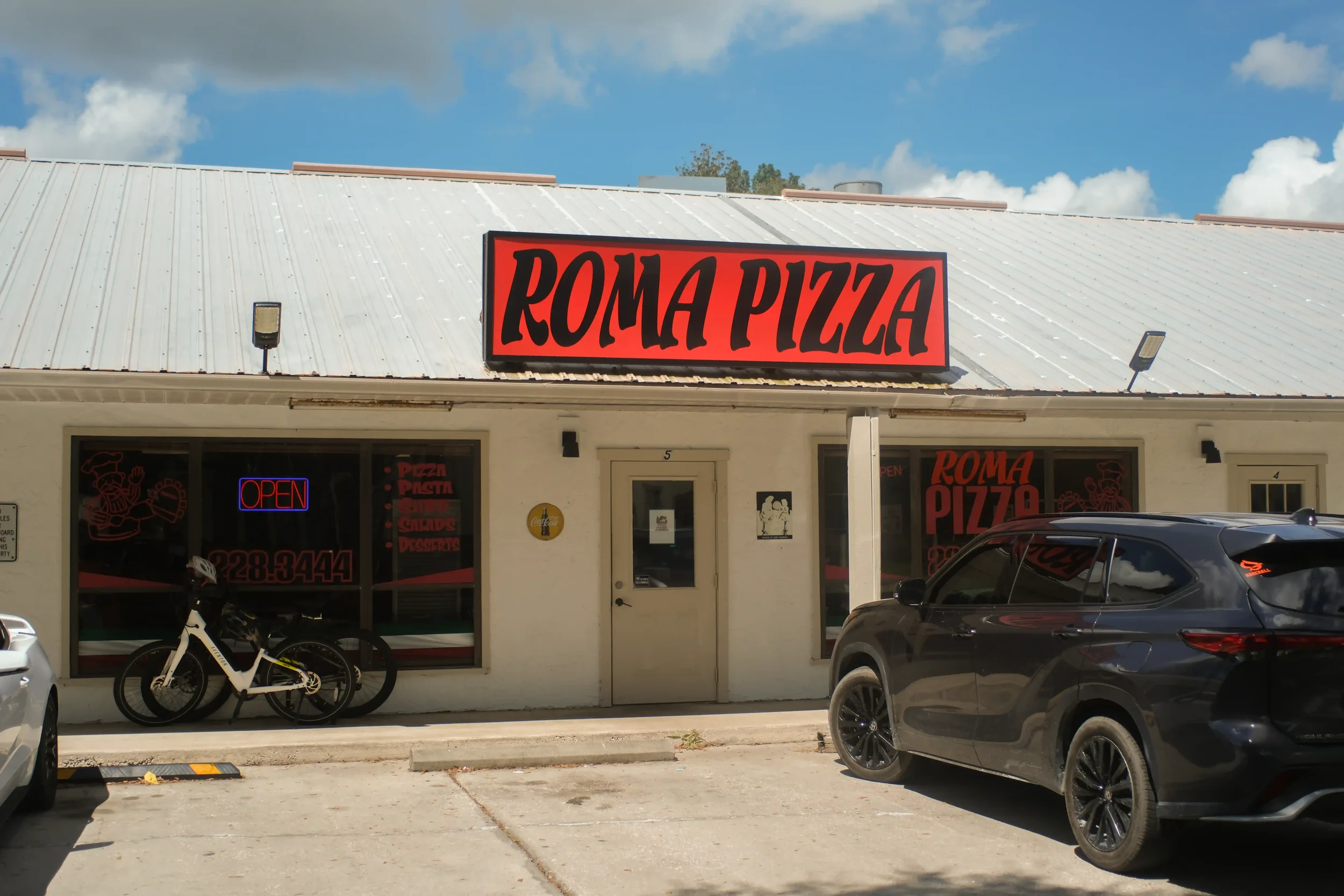 The exterior of the Roma Pizza building featuring a bright red illuminated sign on the roof and vehicles parked out front.