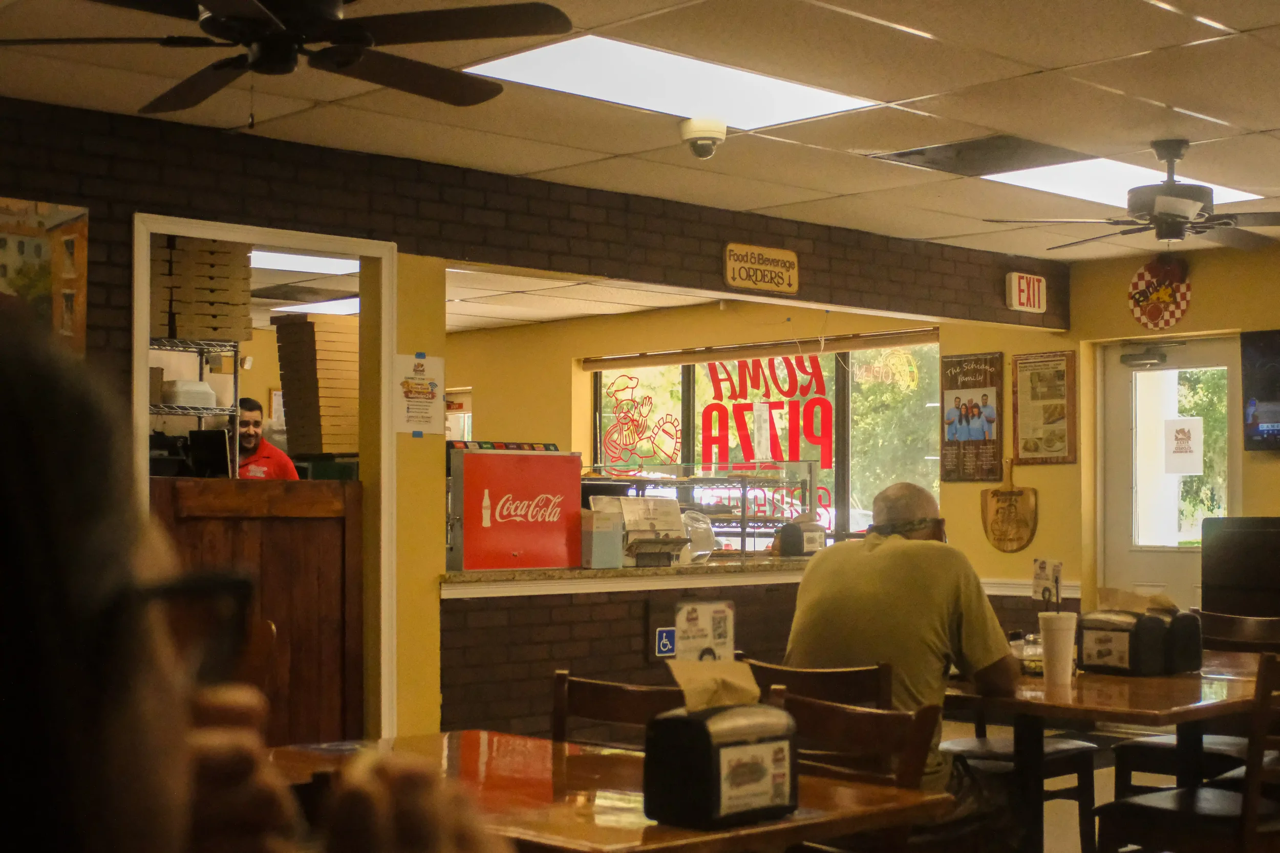 The casual dining room of Roma Pizza with patrons sitting at wooden tables, yellow walls, and a staff member behind the ordering counter.