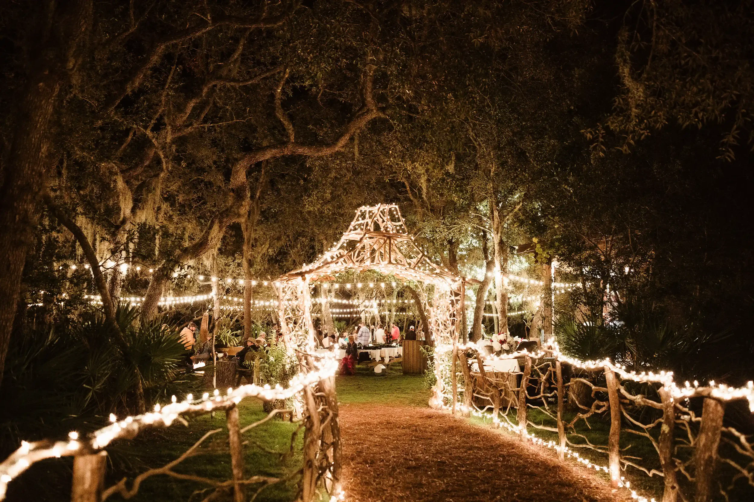 An outdoor event space at night featuring a rustic wooden archway and a pathway illuminated by warm string lights with guests gathered at tables in a wooded background.