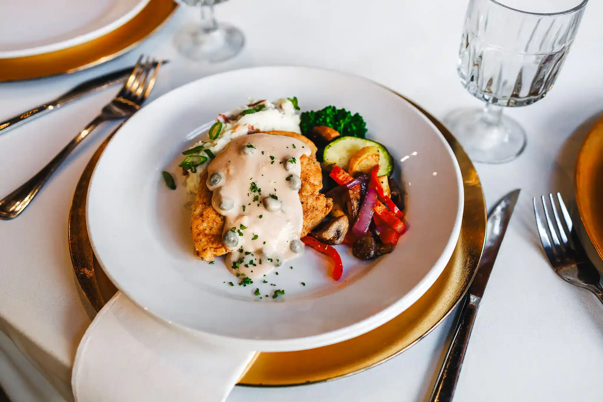 A plated dinner featuring chicken topped with a creamy caper sauce served alongside mashed potatoes and roasted mixed vegetables on a white plate with a gold charger.