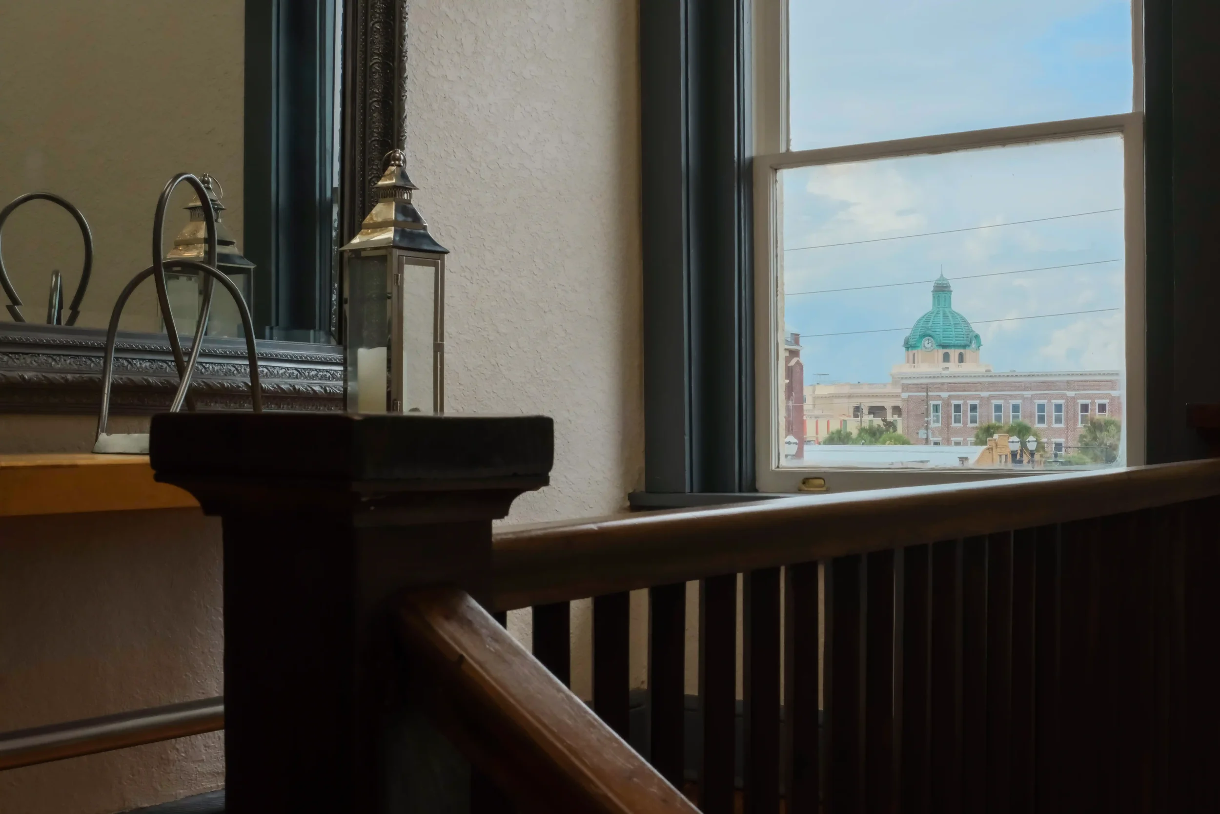 The green dome of the Volusia County Courthouse viewed through a window, positioned behind a wooden staircase railing and a decorative lantern.