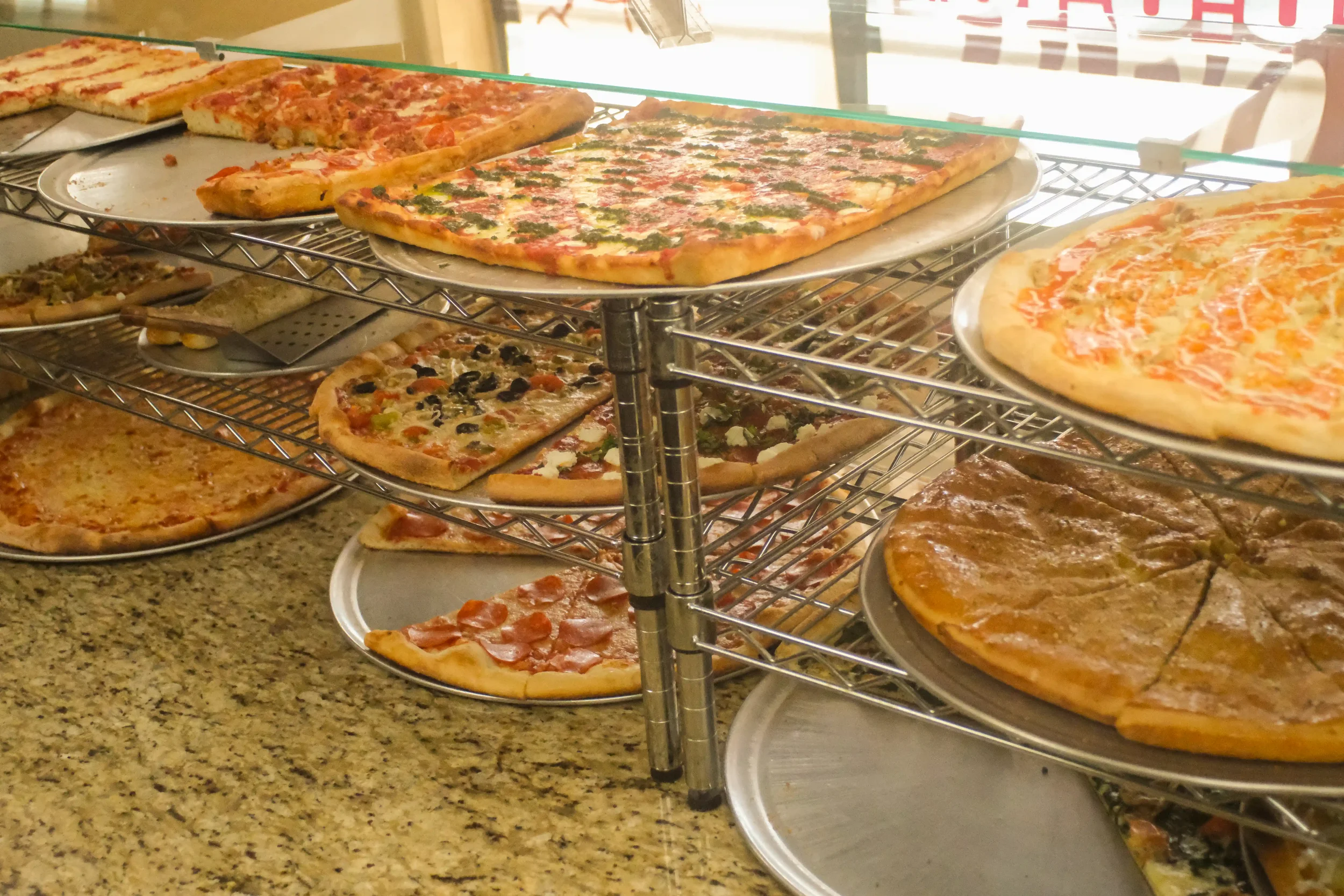 Stacks of cooked New York style pizzas resting on metal racks behind a glass display counter at Roma Pizza.