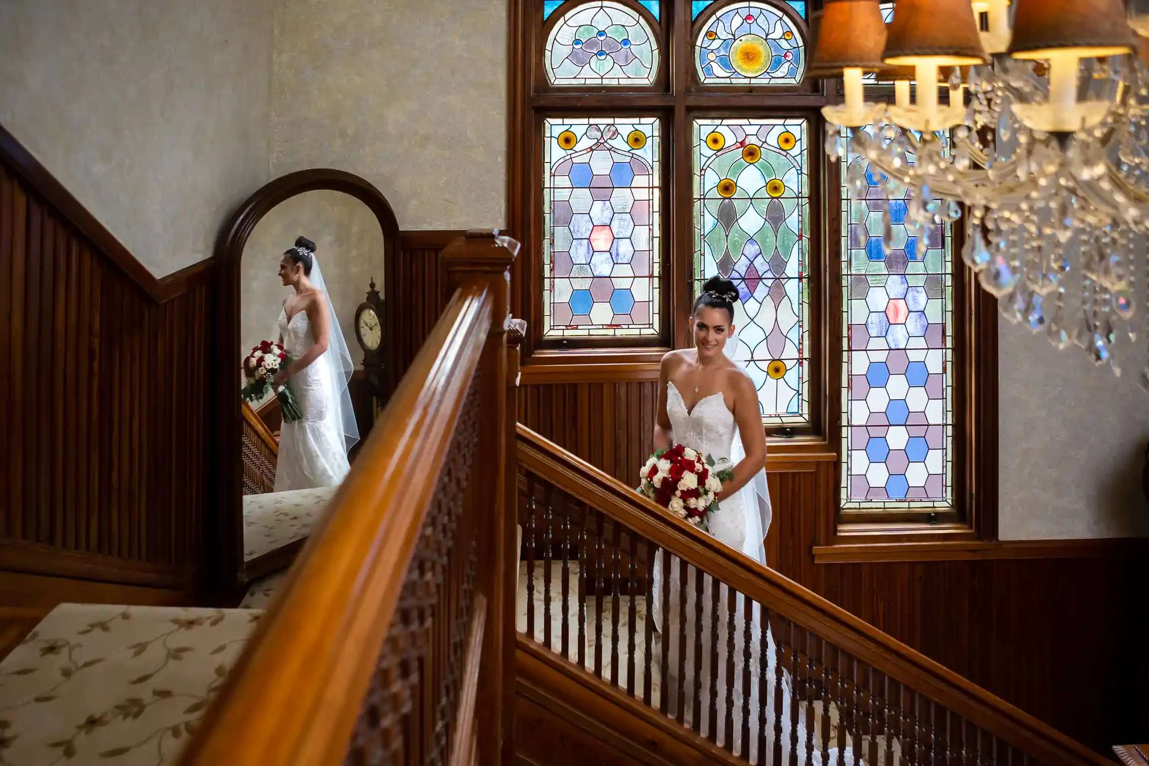 A bride in a strapless white gown holding a red and white rose bouquet standing on an ornate wooden staircase in front of colorful stained glass windows.