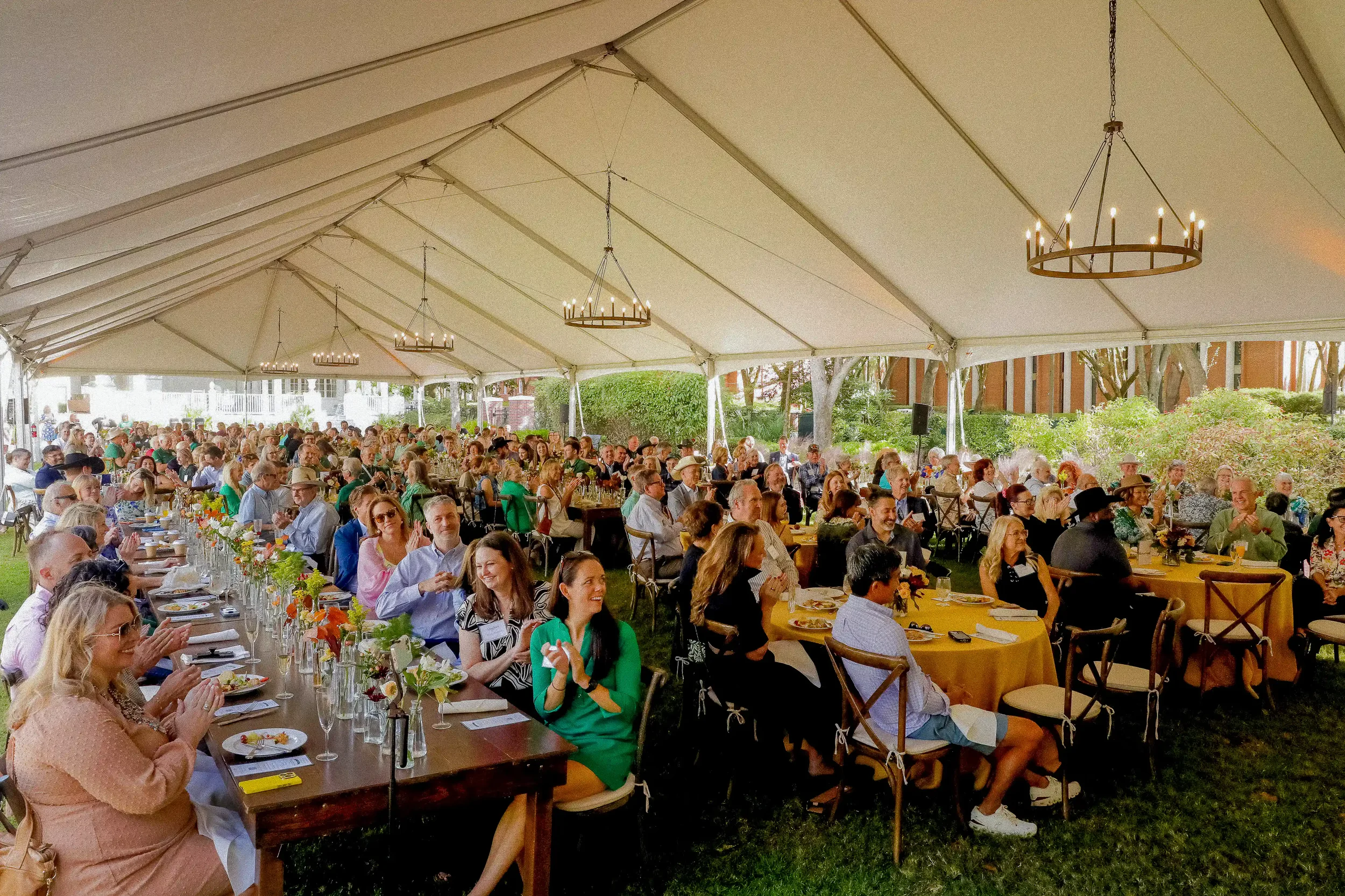 A large group of people dining at long wooden tables and round yellow tables under a spacious white event tent outdoors.