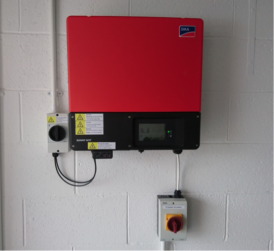 A red solar inverter mounted on a white brick wall with a small digital display, connected to a white electrical box with a red emergency stop button.