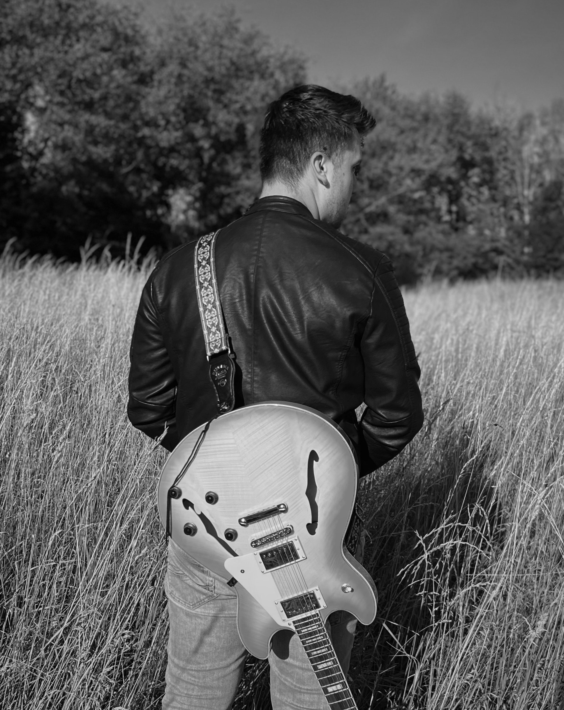 T.R.A.V.L.R wearing a leather jacket with a guitar on his back in a field of tall grass, facing away from the camera, with trees in the background.