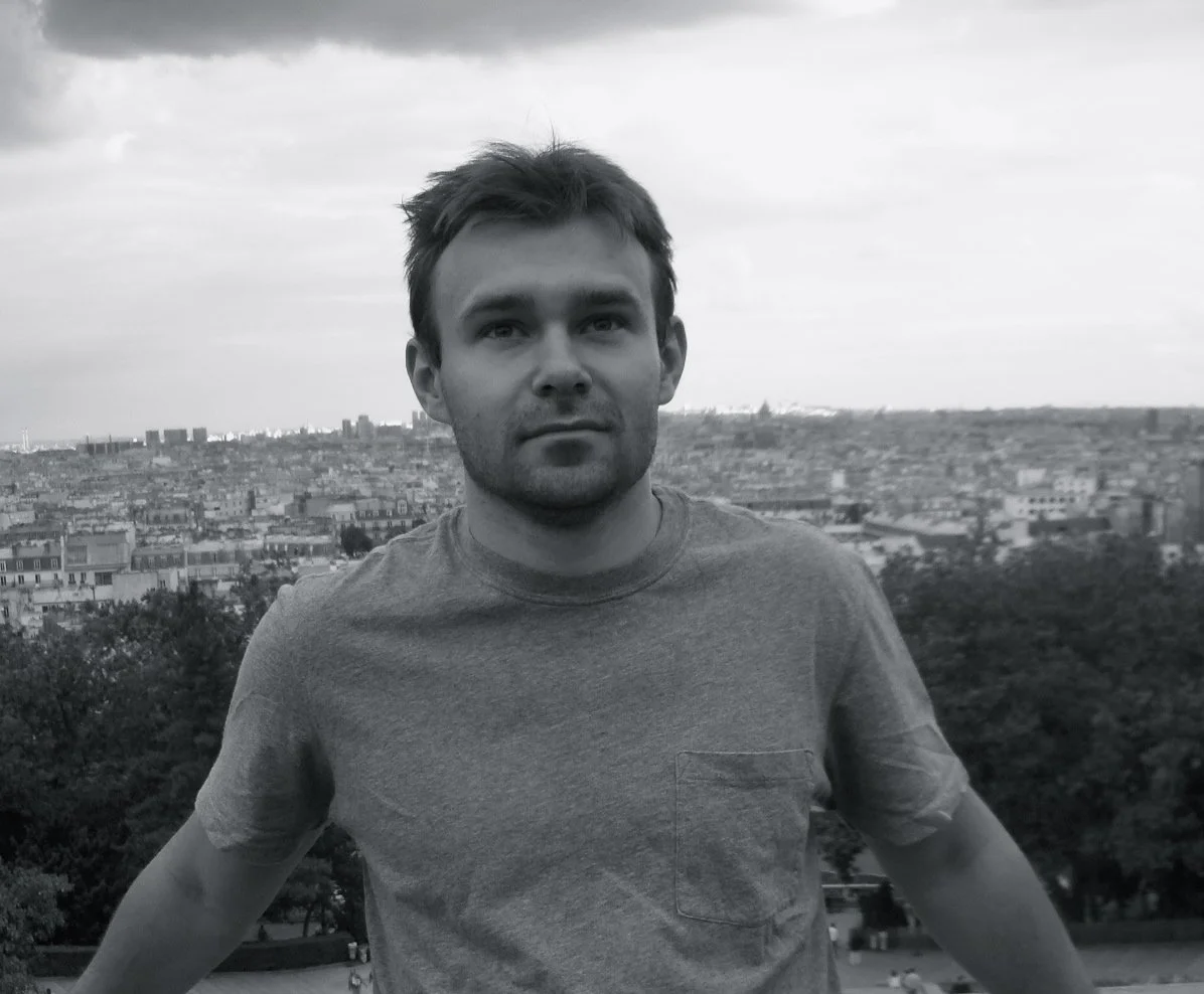 Black and white photo of a young man with short hair, wearing a t-shirt, standing outdoors with a cityscape in the background.