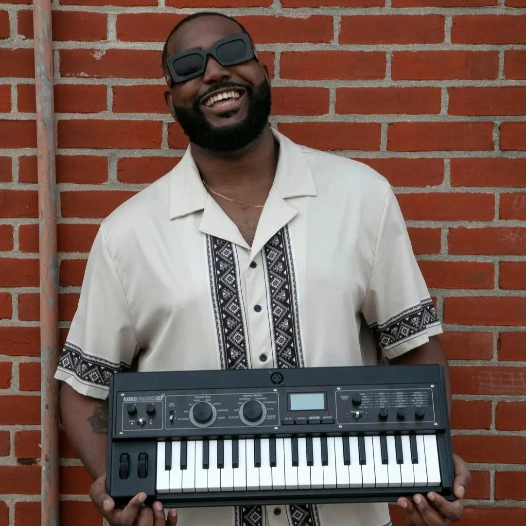 Arthur Luis smiling in sunglasses holding a keyboard synthesizer against a brick wall.