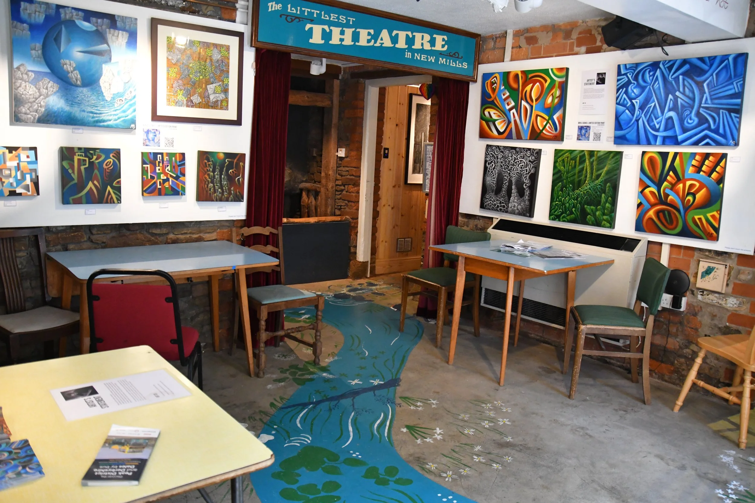 Interior of art gallery with framed landscape paintings on white walls, a woman sitting at a table writing, a brown sofa with pillows, and a chalkboard with handwritten notes.