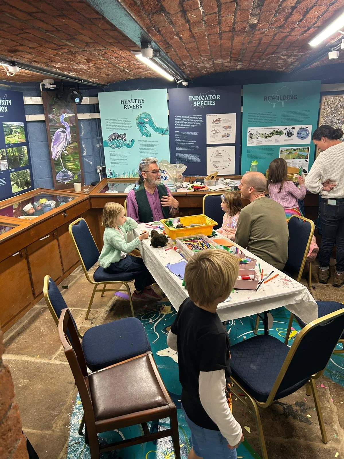 A group of children and adults sit around a table engaging in arts and crafts activities in an indoor exhibit space with educational posters about rivers and wildlife on the walls.