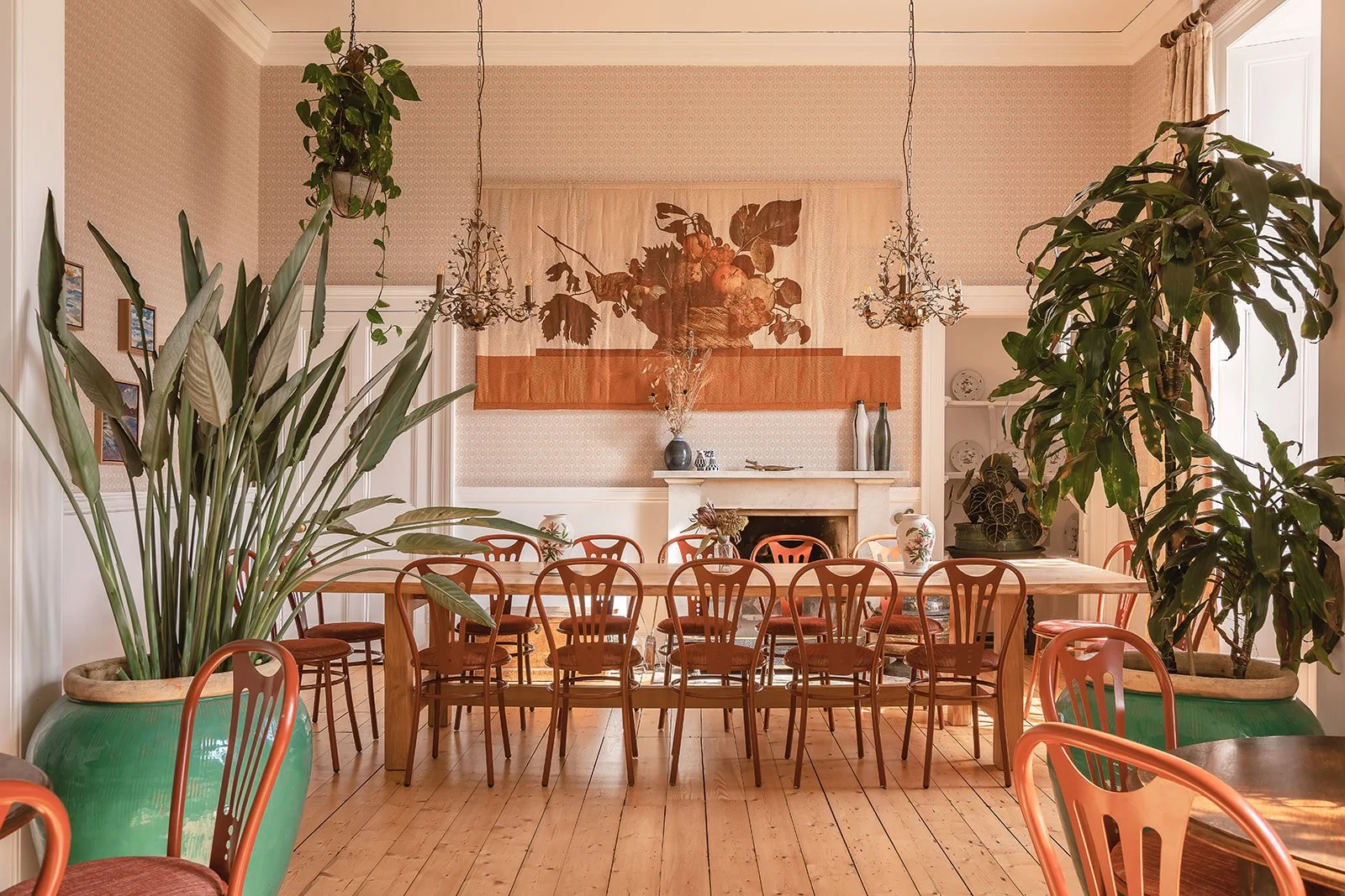 A spacious dining room with wooden floors and large potted plants, featuring a long wooden table surrounded by wooden chairs, with a fireplace and decorative artwork on the wall in the background.