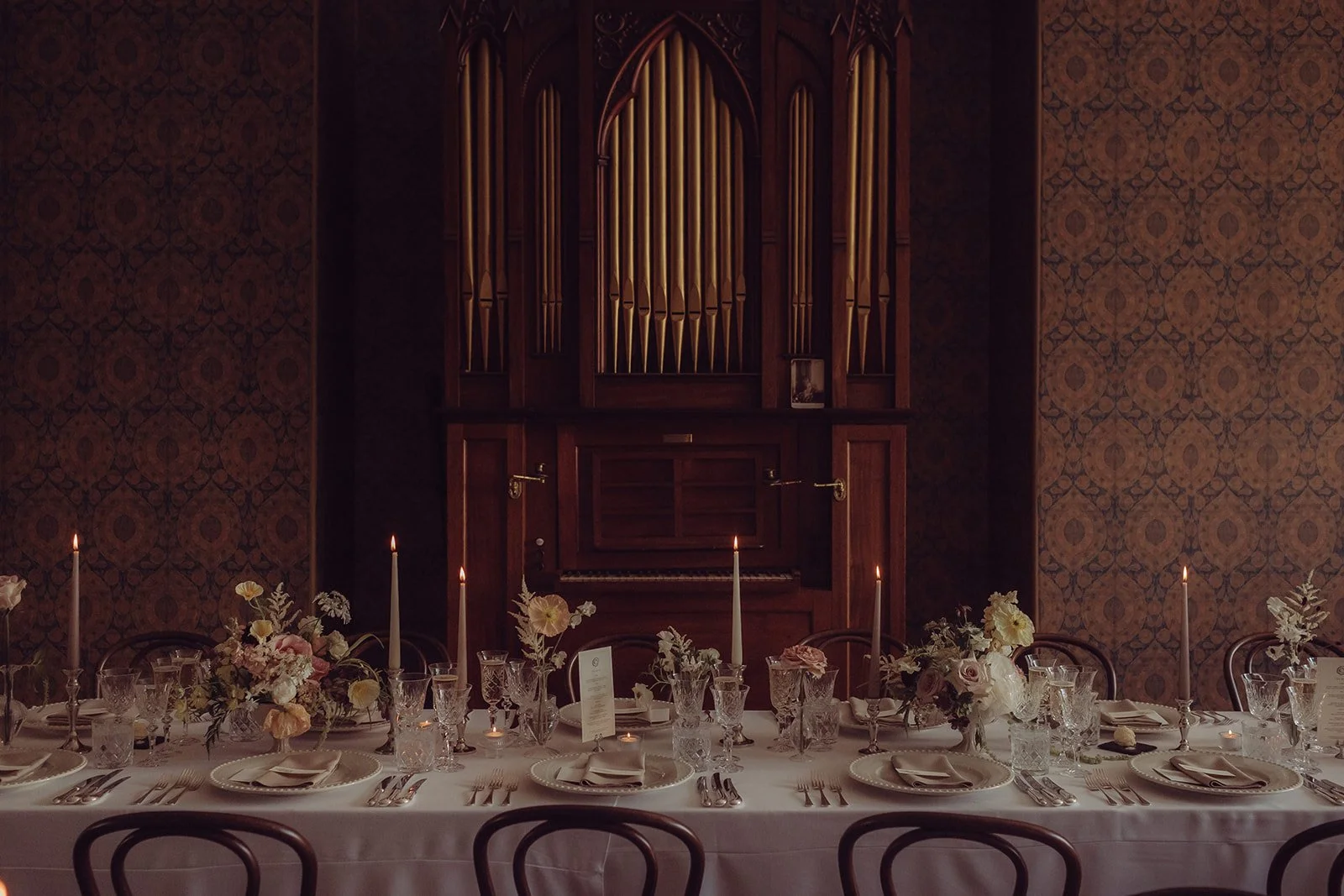 Elegant dining room with a long table set with glassware, silverware, and candles, with a large pipe organ against a patterned wall in the background.