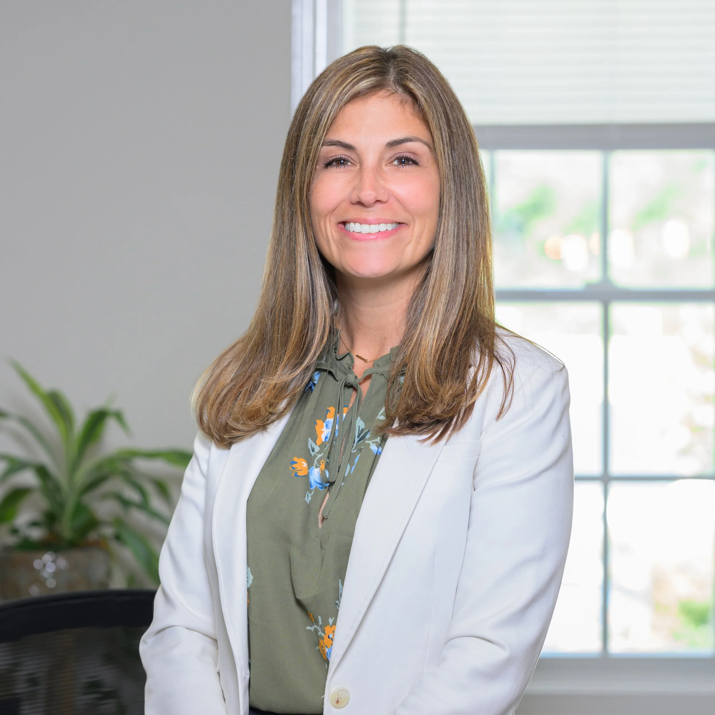 A woman with long brown hair, wearing a white blazer over a green blouse with a floral pattern, smiling in an indoor setting with a window and a plant in the background.