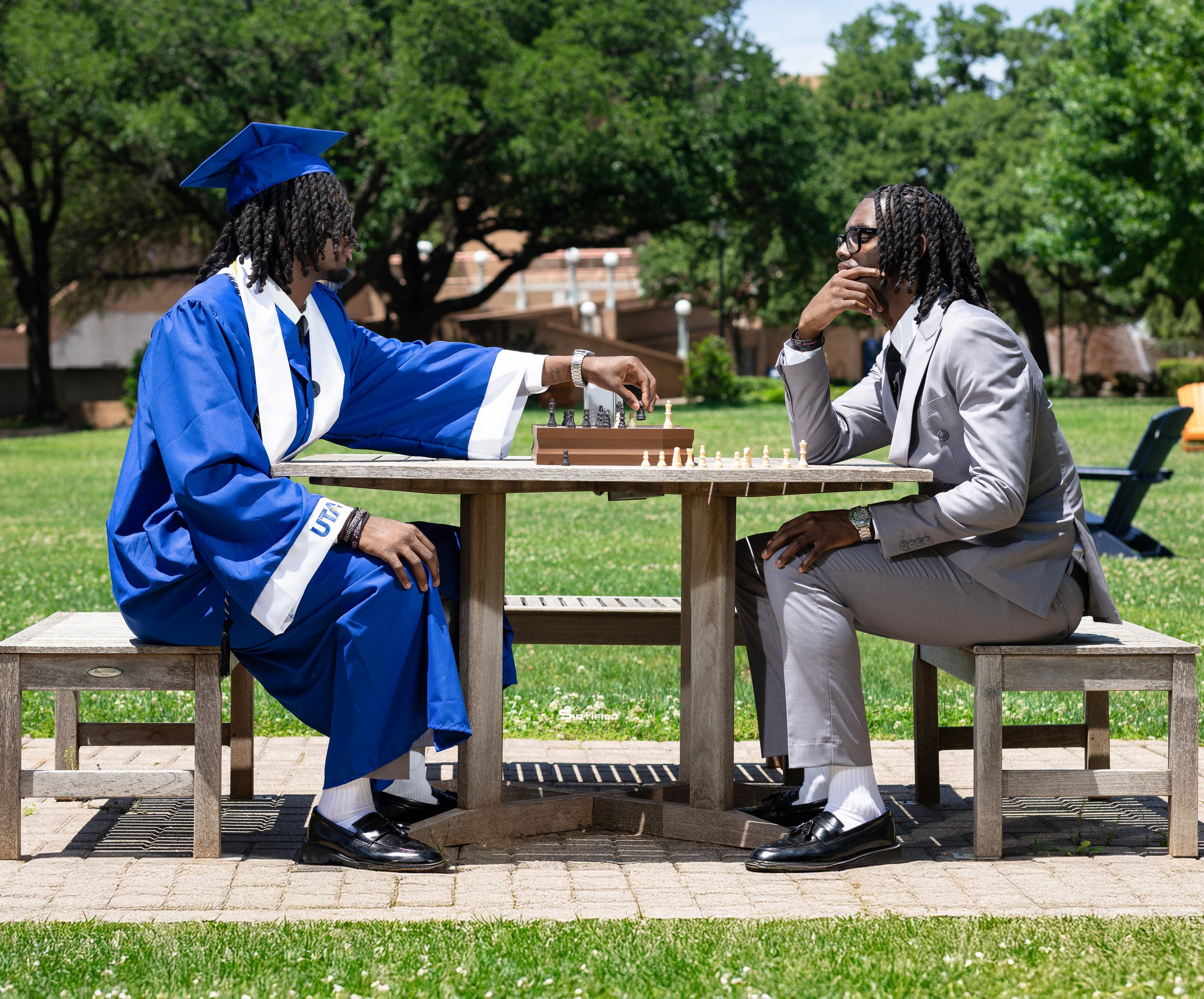 A person in a blue graduation gown and cap playing chess with another person in a light grey suit outdoors on a park bench.