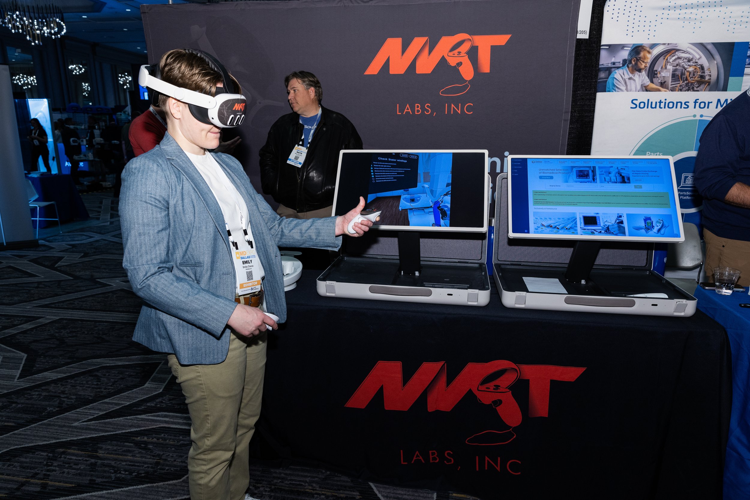 A woman wearing a virtual reality headset and holding a controller, standing in front of two display screens at a booth for NMT Labs, Inc. Two men are seen in the background talking, and a banner with the company's logo is behind the booth.
