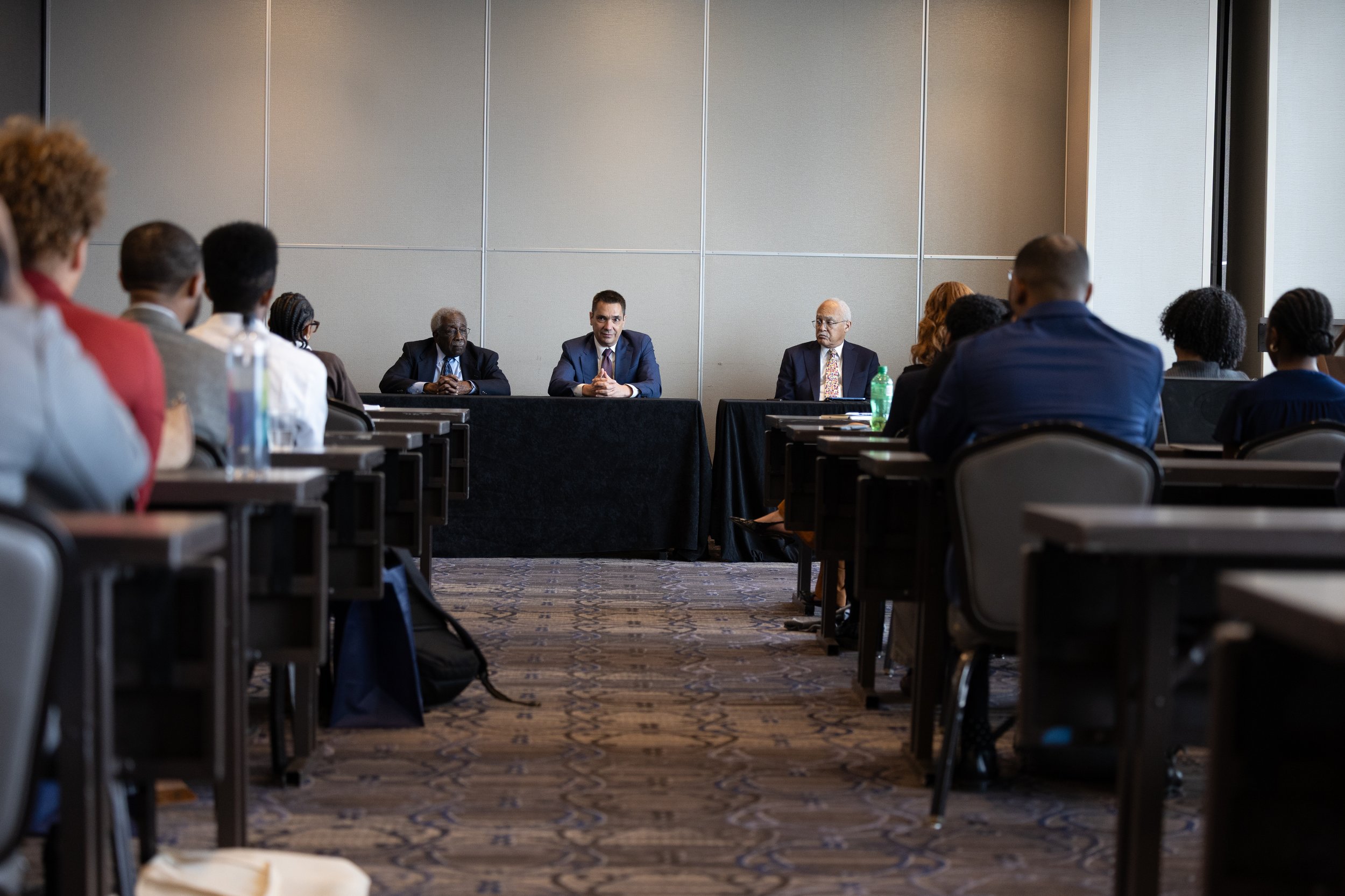 A conference room with a panel of three men at the front and an audience seated in rows. The panelists are dressed in suits, and the room is well-lit with beige walls.