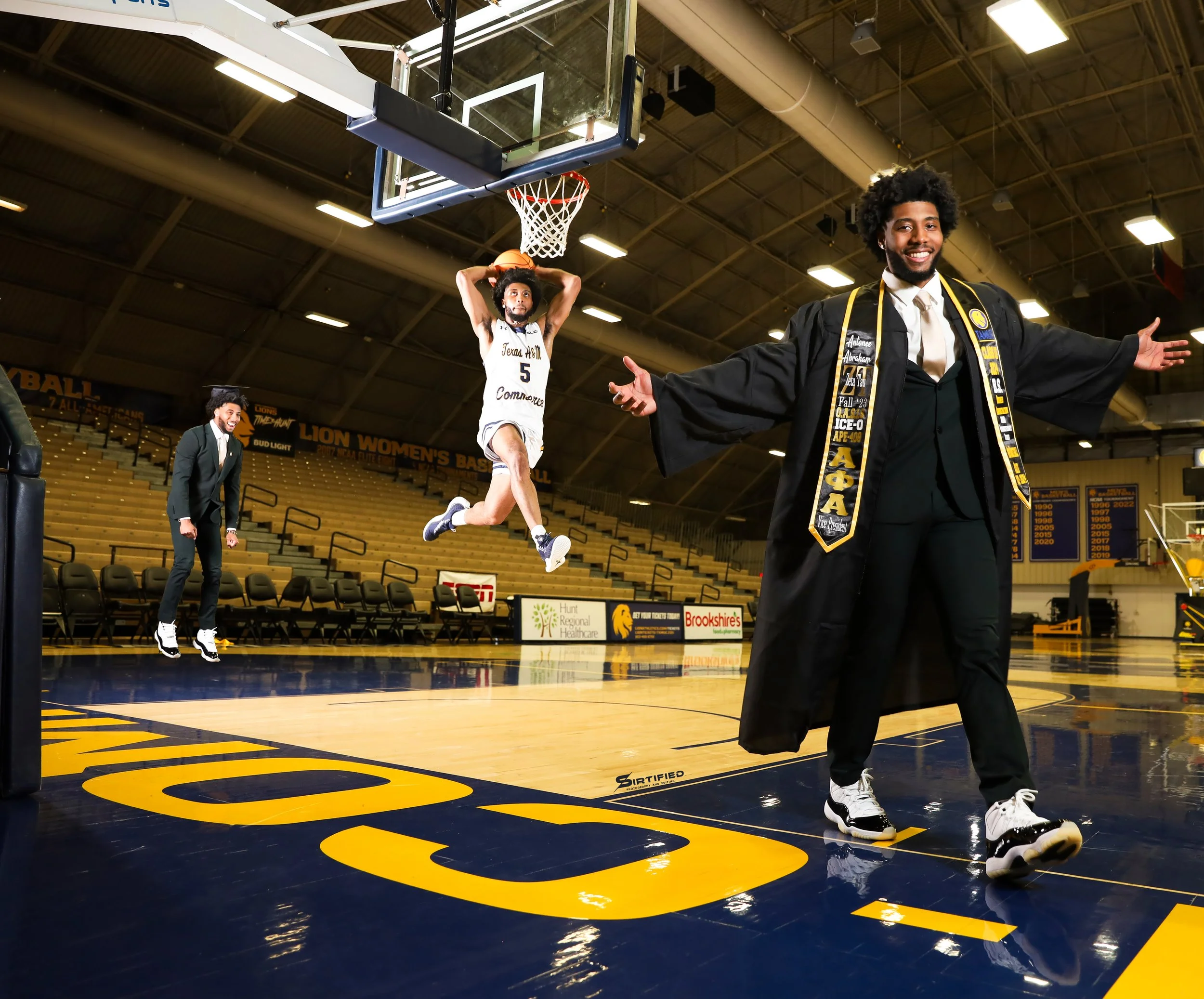 Basketball player in a white jersey jumping to dunk the ball near the hoop, with a man in academic regalia celebrating on the court, while another man in a suit watches in the background. The court has yellow markings and is in an indoor arena.
