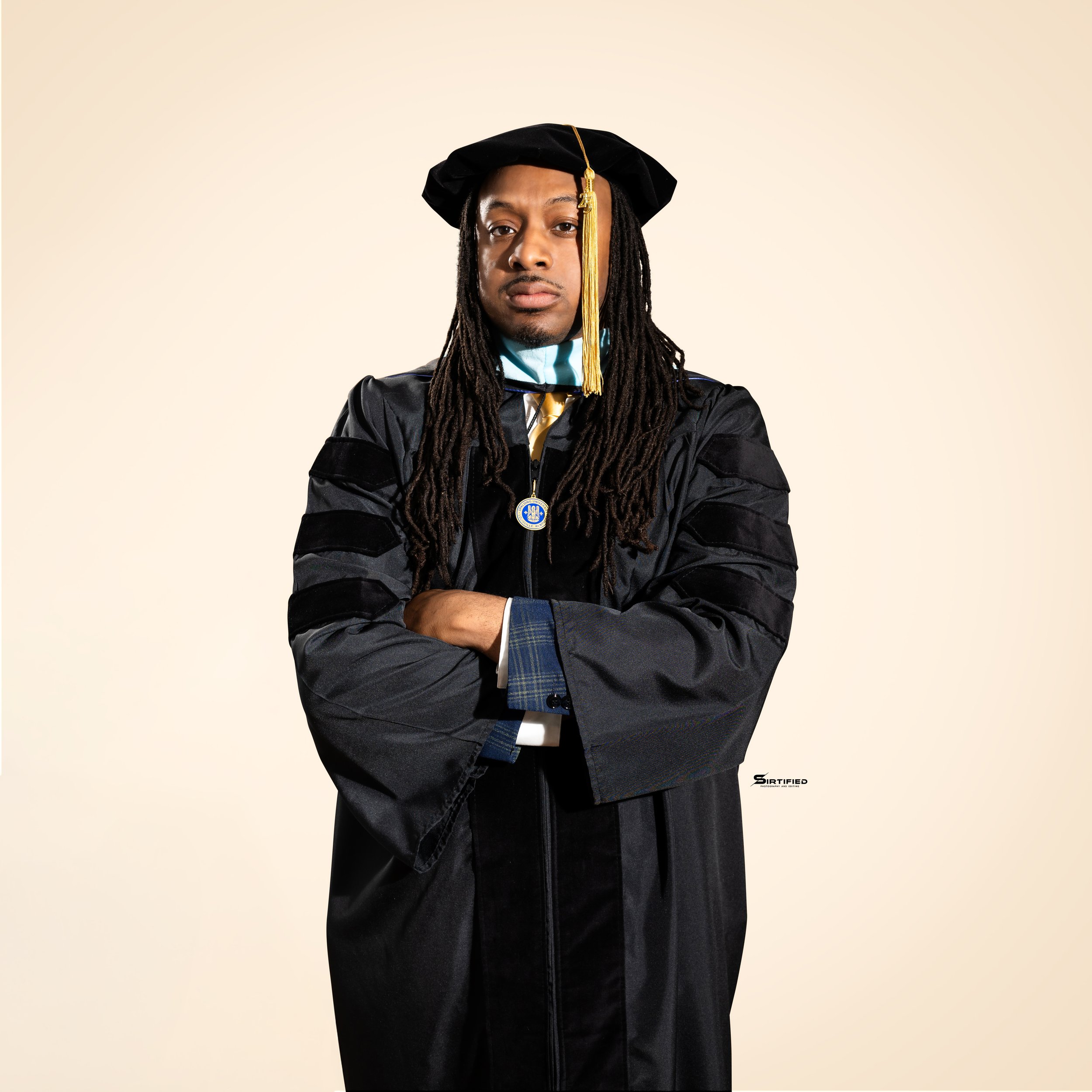A person in academic regalia with a black gown and cap, gold tassel, and medallion, standing with arms crossed against a plain background.