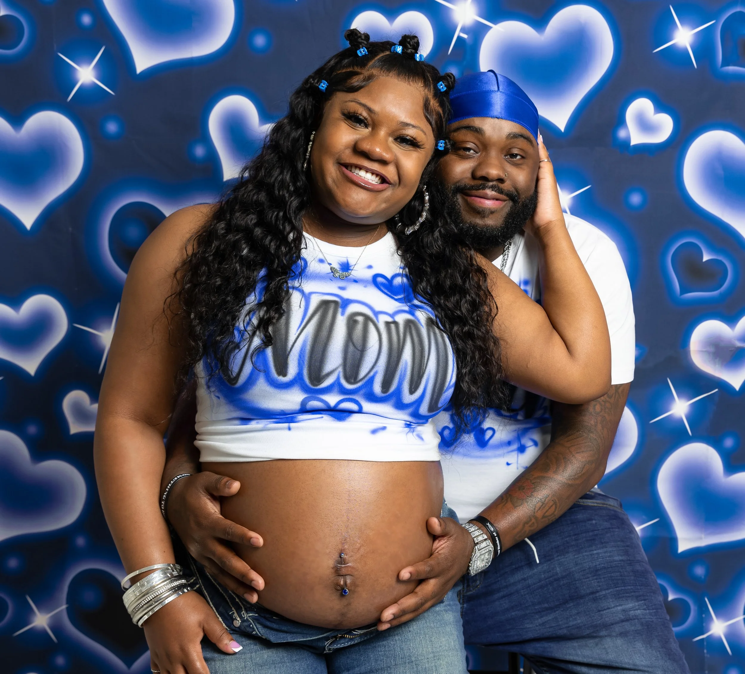 A pregnant woman and her partner smiling and posing together against a blue heart-themed background. The woman has long, curly hair and wears a white crop top with blue graffiti-style writing. The man has a beard and is wearing a blue durag and a whi
