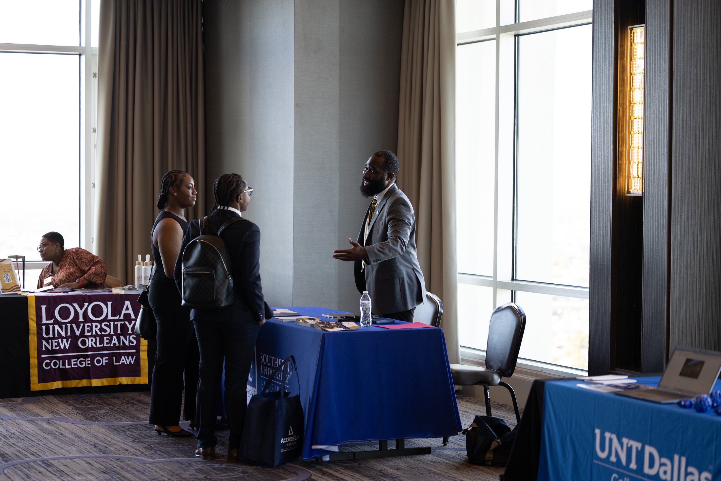 At a career fair, two women and a man are engaged in conversation. The women are wearing business attire, one with a backpack, and the man is in a suit. The booth displays banners for Loyola University New Orleans College of Law and UNT Dallas.