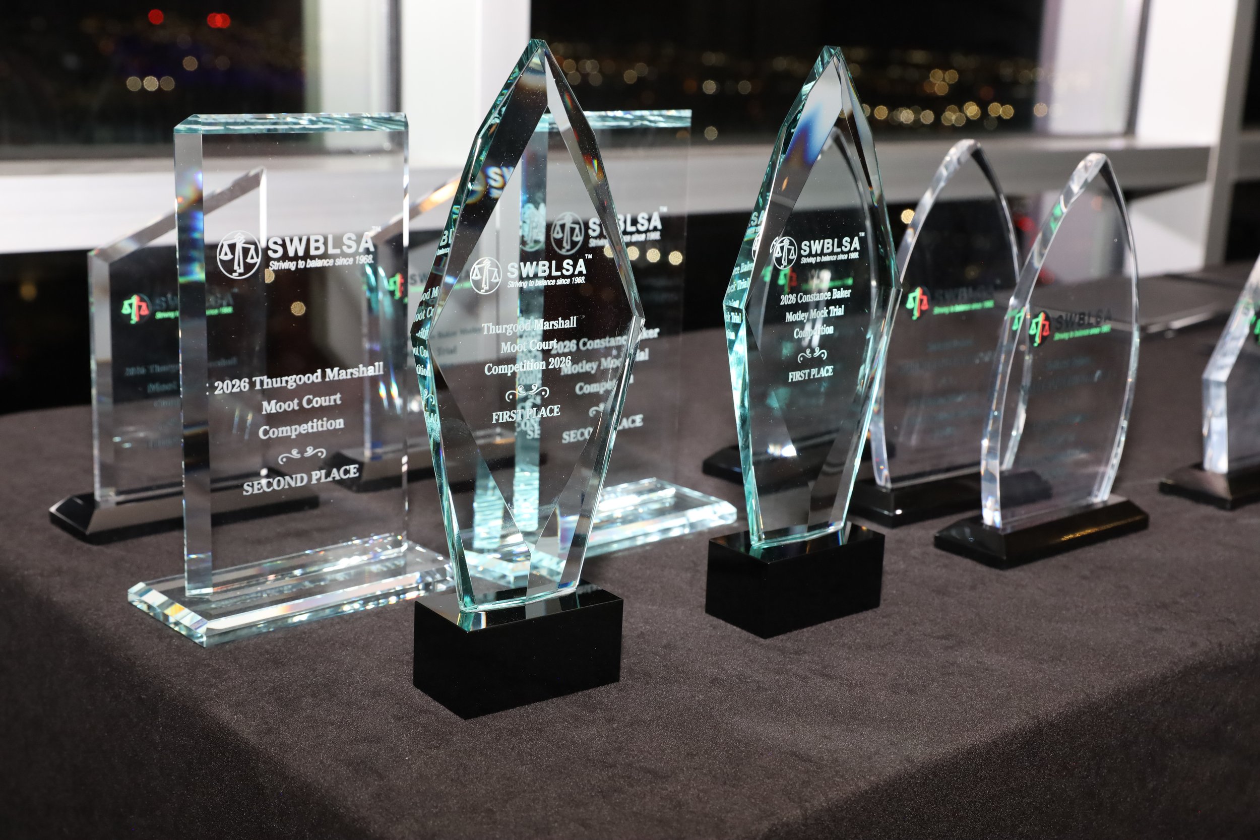 Multiple glass trophies with engraved text, displayed on a dark table in front of a window at night.