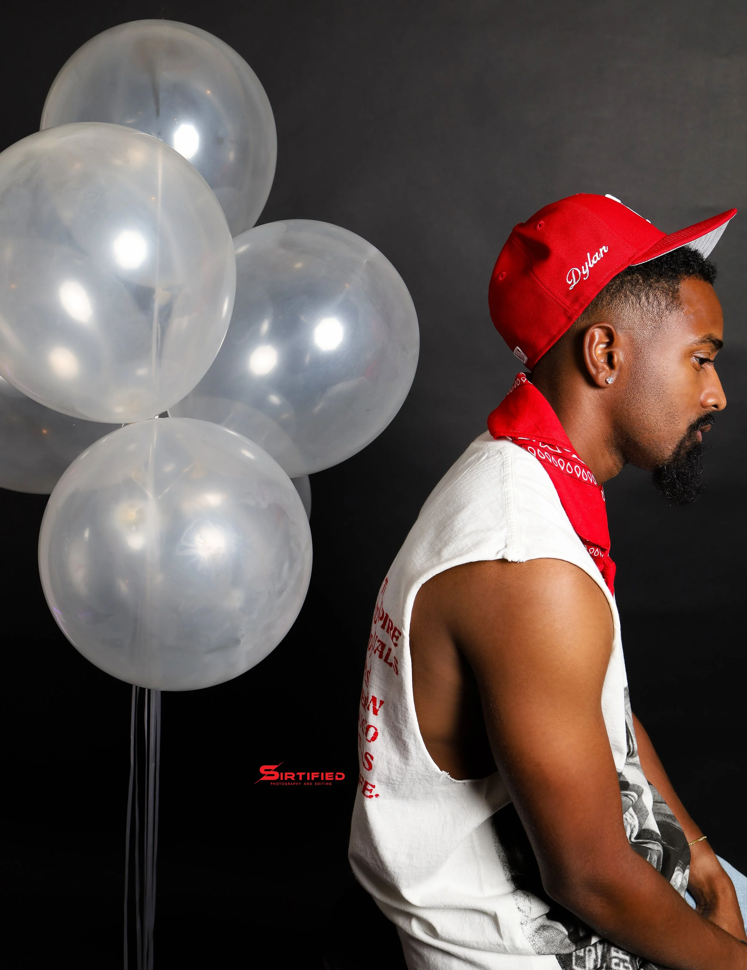 A young man with a beard wearing a red baseball cap with "Dylan" embroidered on it, a red bandana around his neck, and a sleeveless white shirt sitting against a dark background with white balloons to his left.