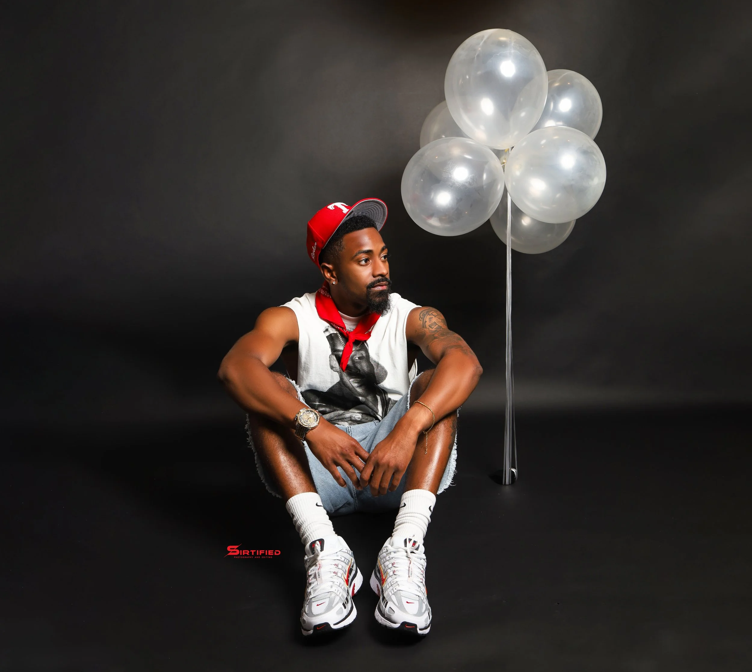 A man sitting on the floor with his legs crossed, wearing white sneakers, socks, and casual clothing, next to a bunch of white balloons against a dark backdrop.