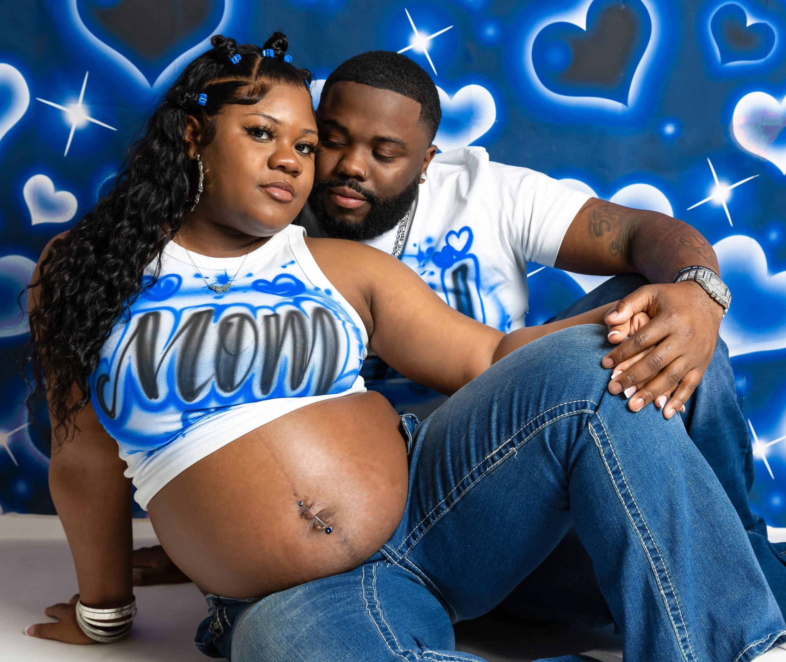 A couple sitting closely, the woman is pregnant with a belly piercing, and they are in front of a blue, heart-themed background.