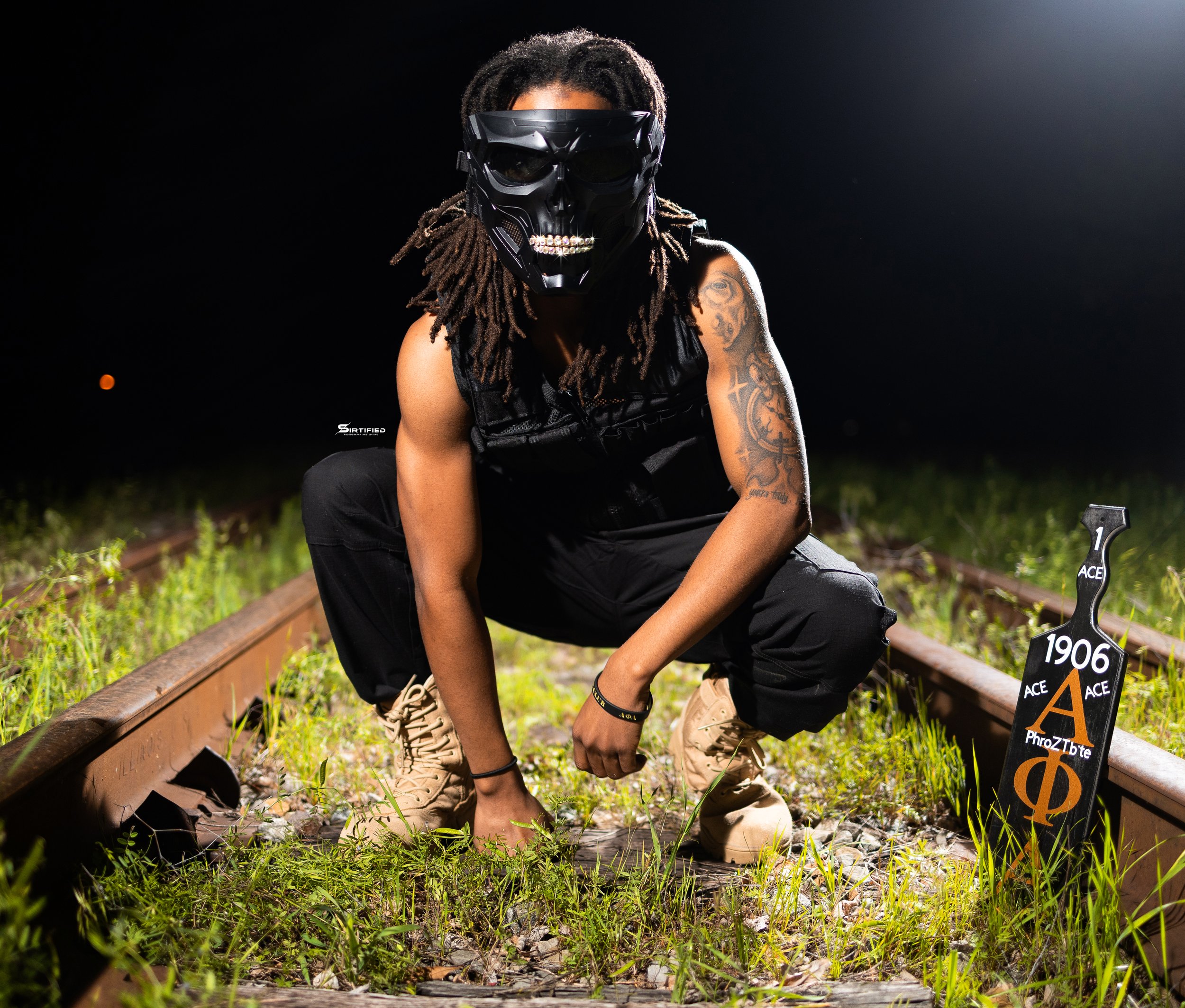 Person with dreadlocks crouching on railroad tracks at night, wearing a skull mask and black clothing, illuminated by a bright light, with a black paddle with Greek symbols and the year 1906 standing nearby, and grass growing on the tracks.