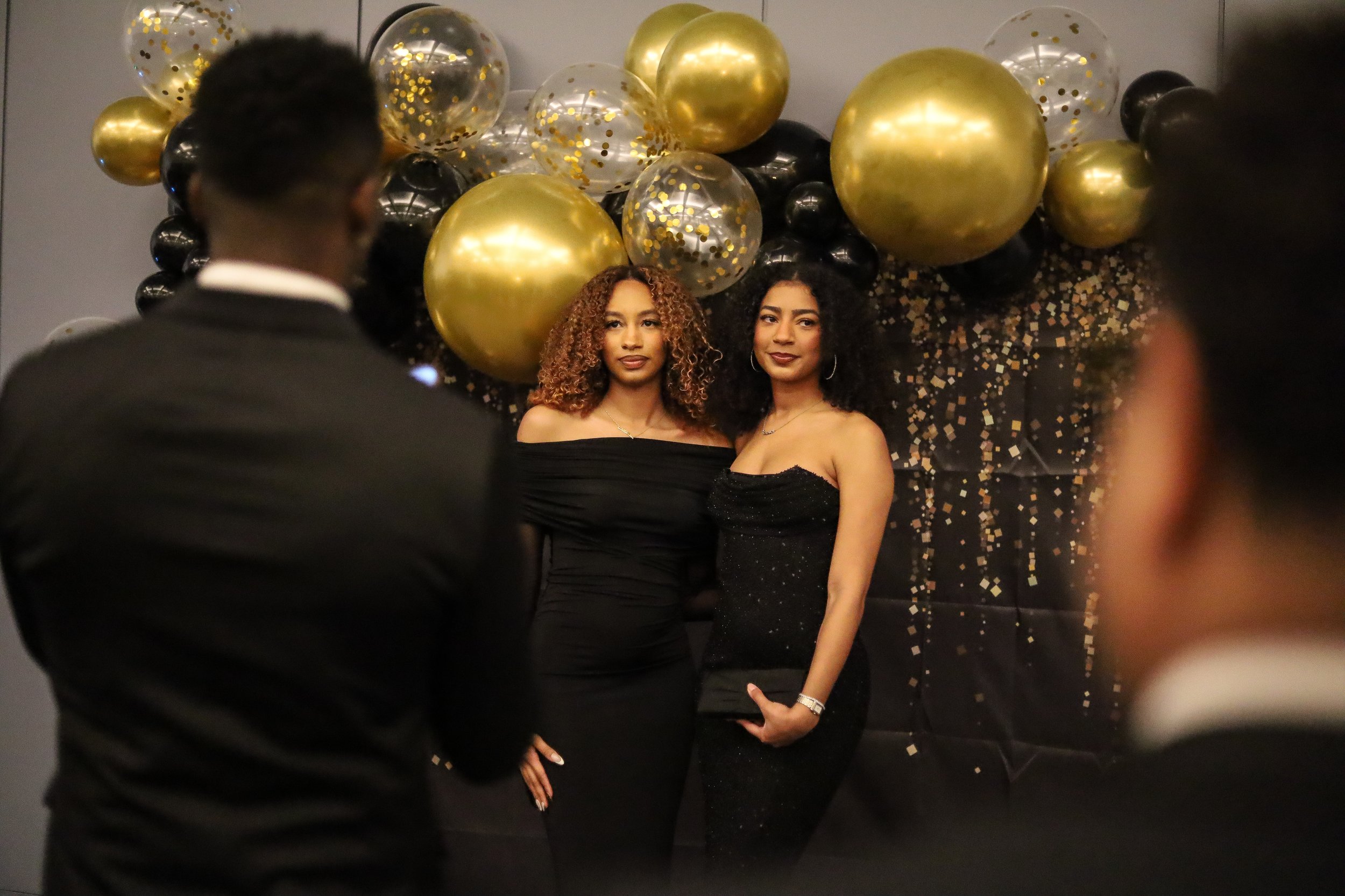 Two women in black dresses posing for photos at a celebration with gold, black, and clear balloons and confetti backdrop, while two men in suits stand in front observing.
