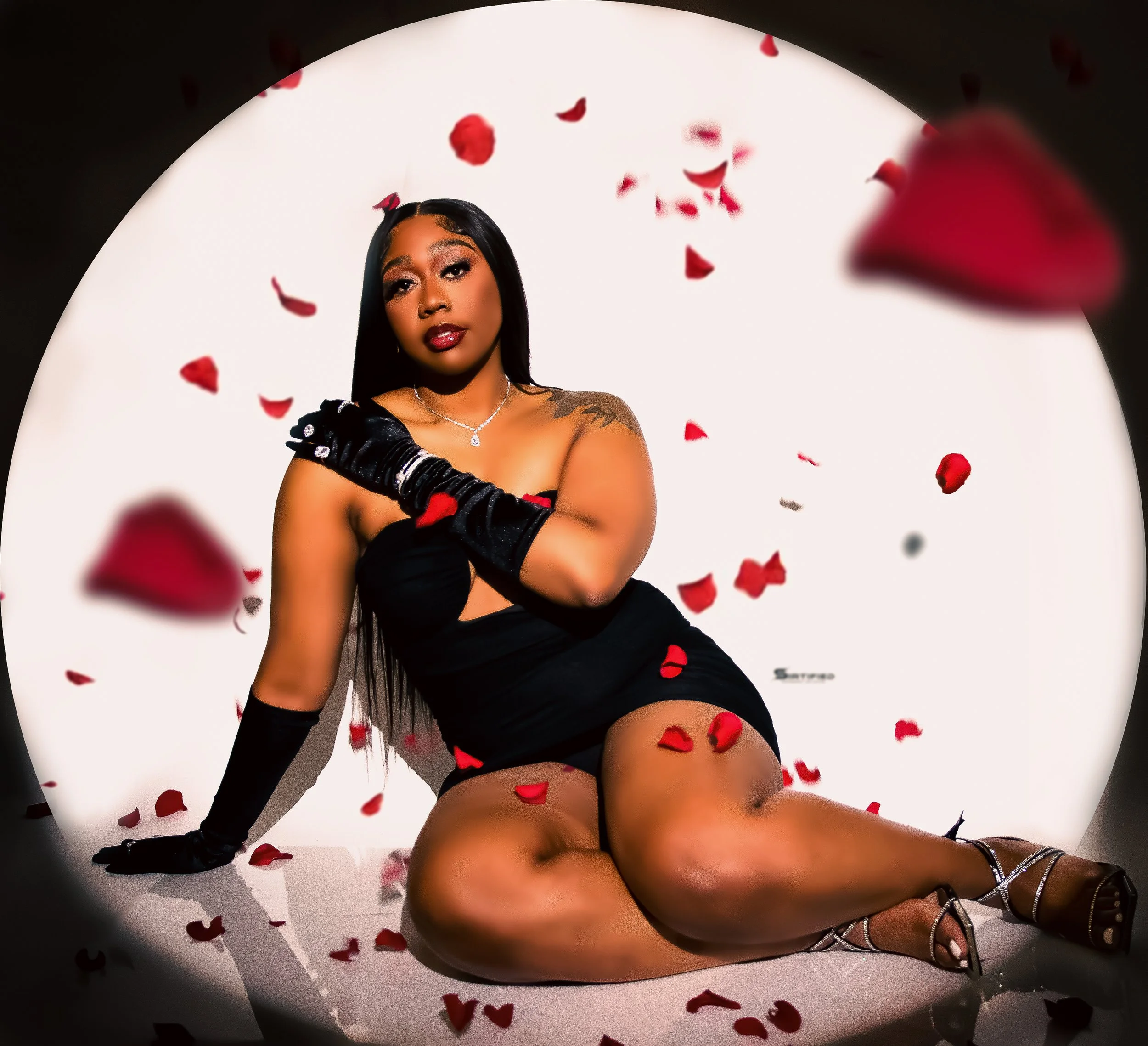 A woman in a black dress sits on the floor surrounded by red rose petals, wearing long black gloves, high heels, and a necklace, with a white circular backdrop.