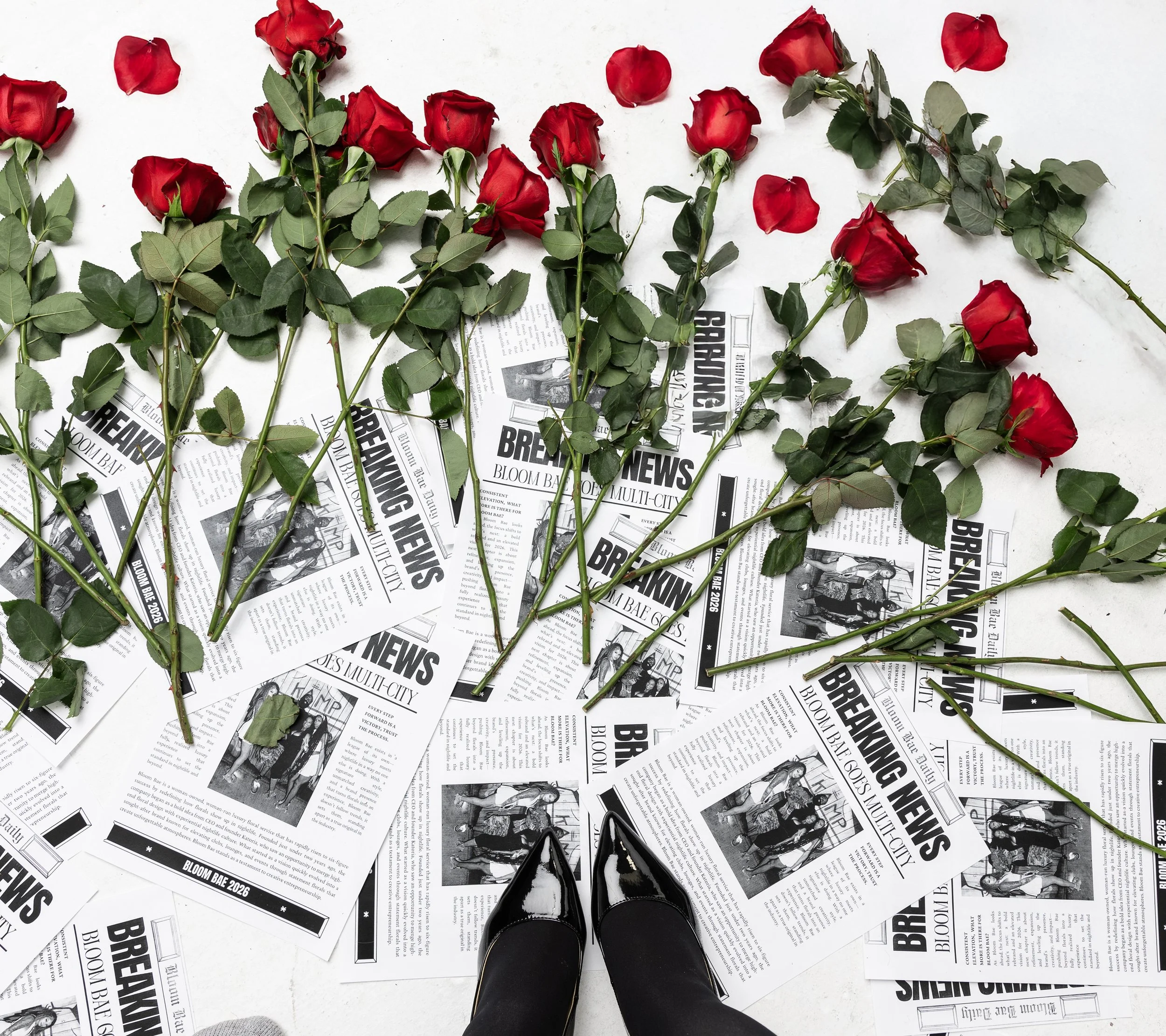 Multiple red roses with green leaves placed on scattered black and white newspapers on a white surface, with pointed black shoes at the bottom.