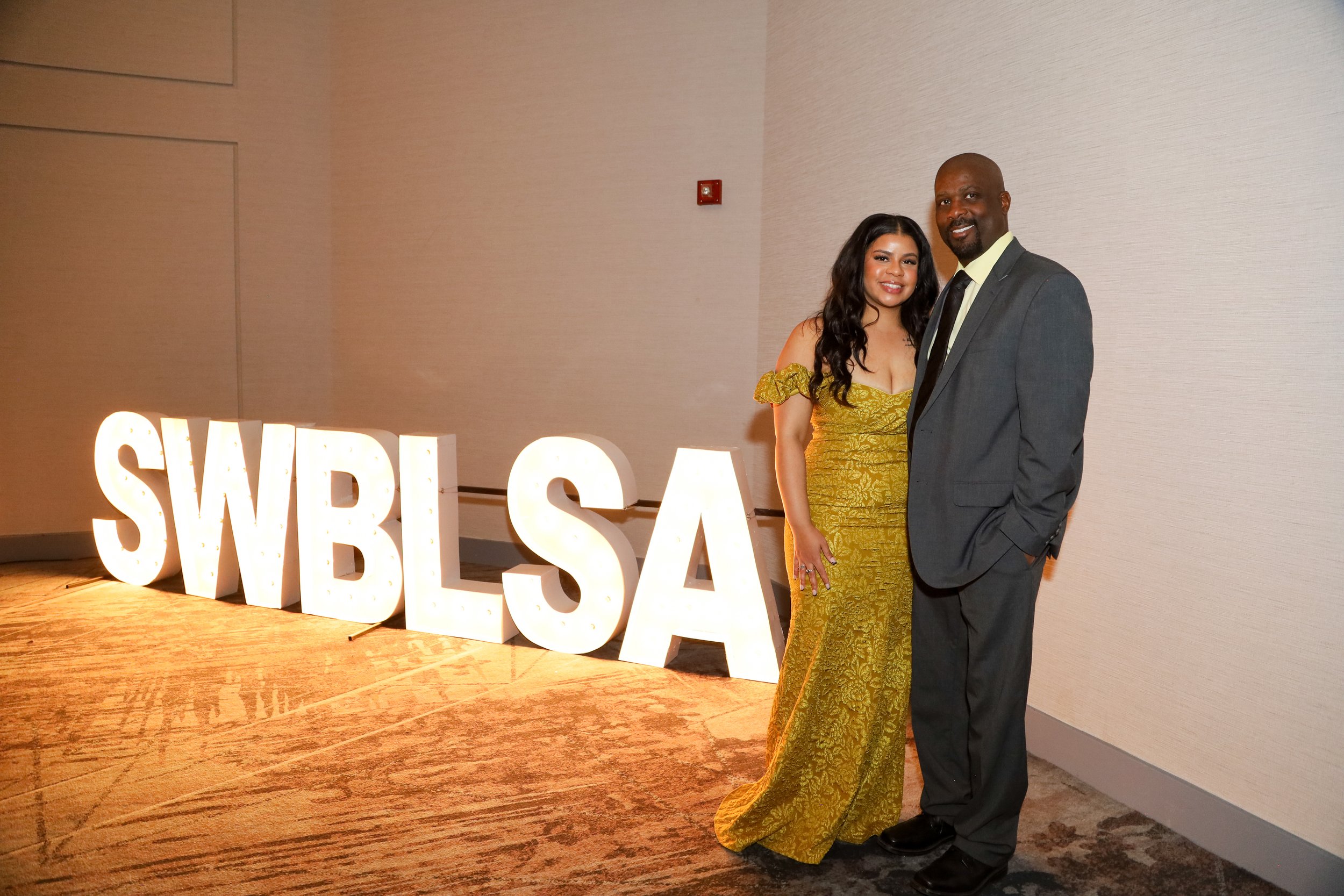 A woman in a yellow gown and a man in a gray suit pose together at a formal event, standing beside large illuminated letters spelling 'SWBLSA'.
