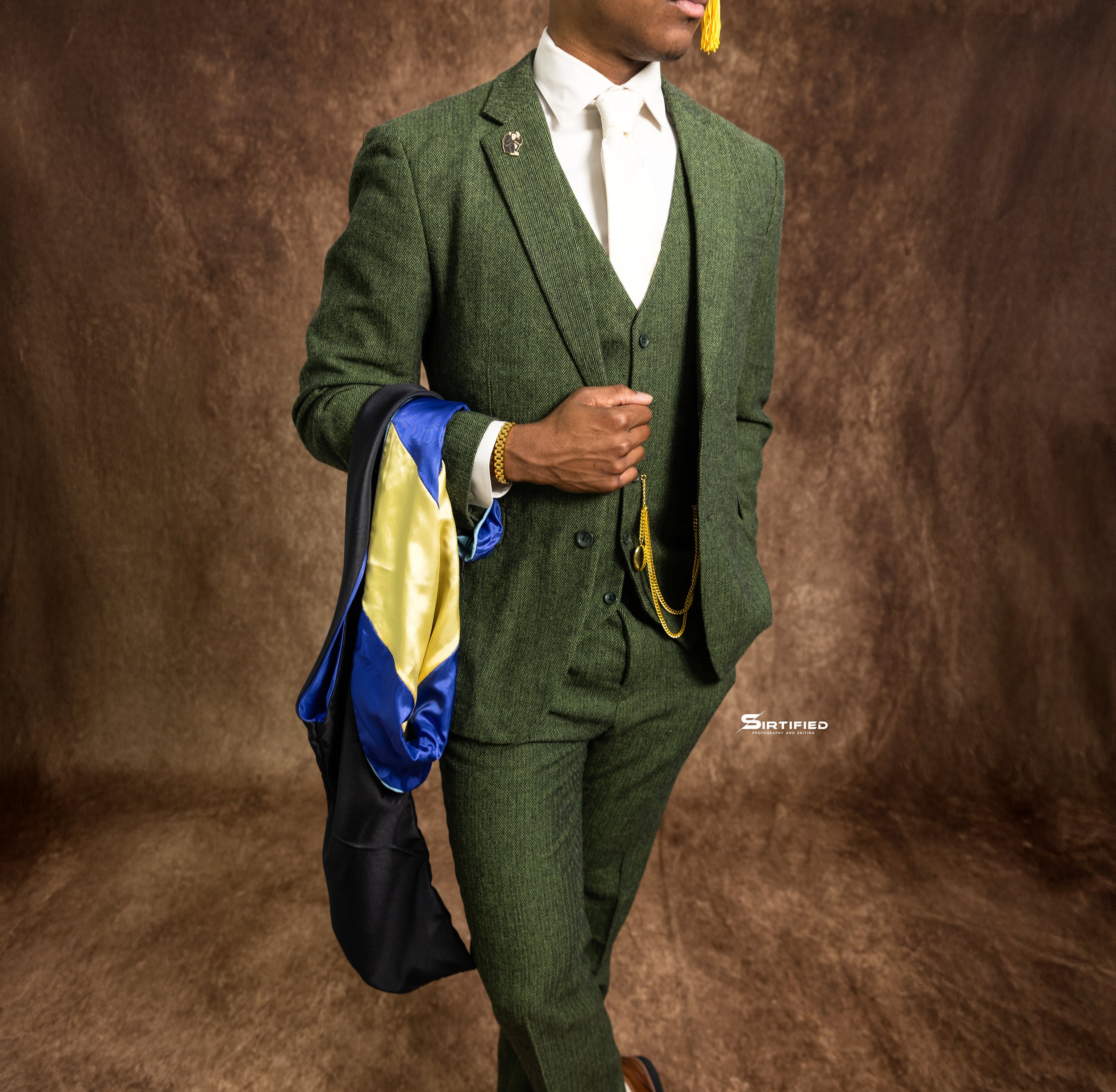 Man dressed in a green suit with a white shirt, holding a colorful jacket, standing in front of a brown backdrop.