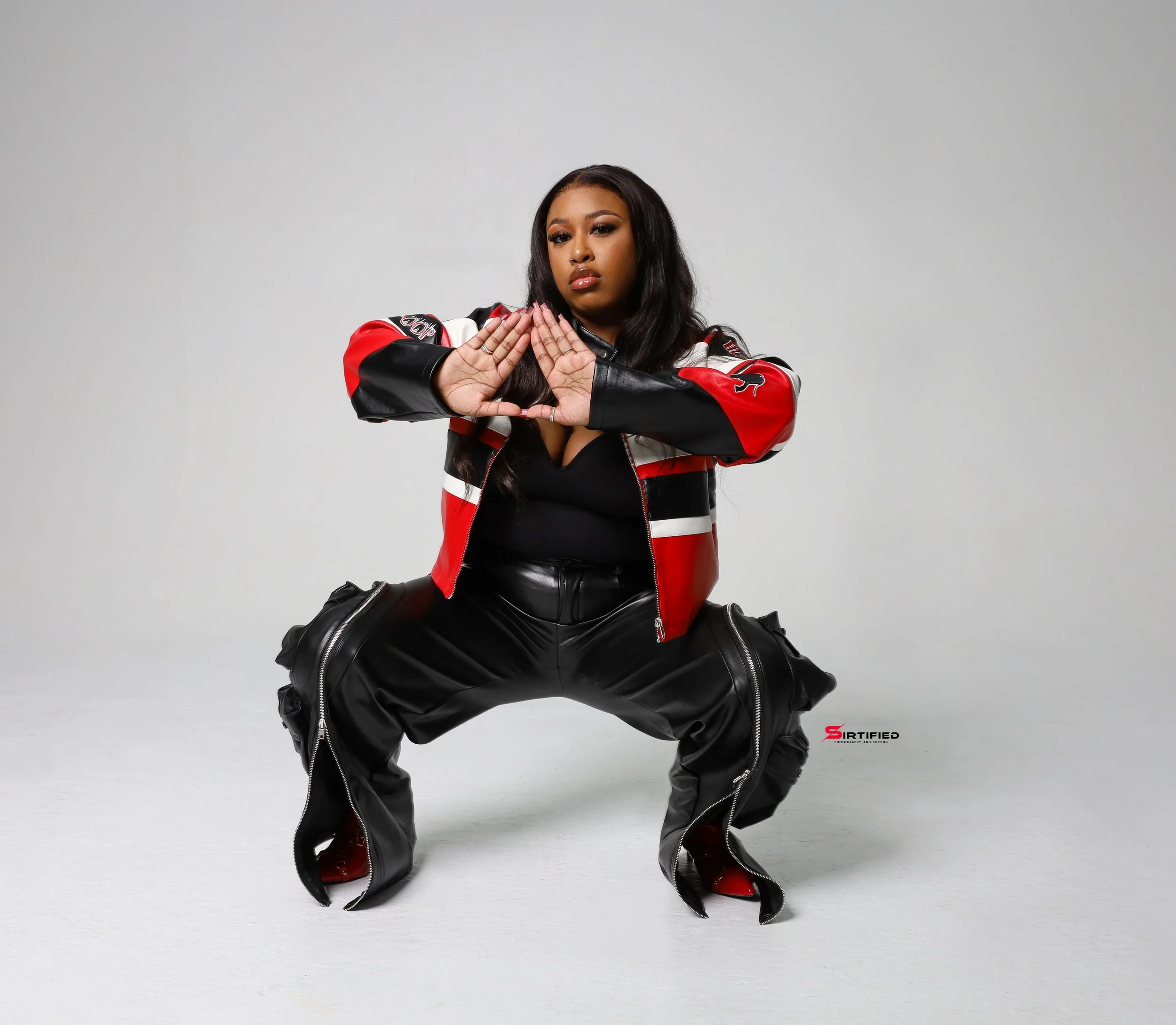 Woman wearing a black shirt, red and black jacket, and black leather pants, squatting with hands together in a prayer position against a plain light-colored background.