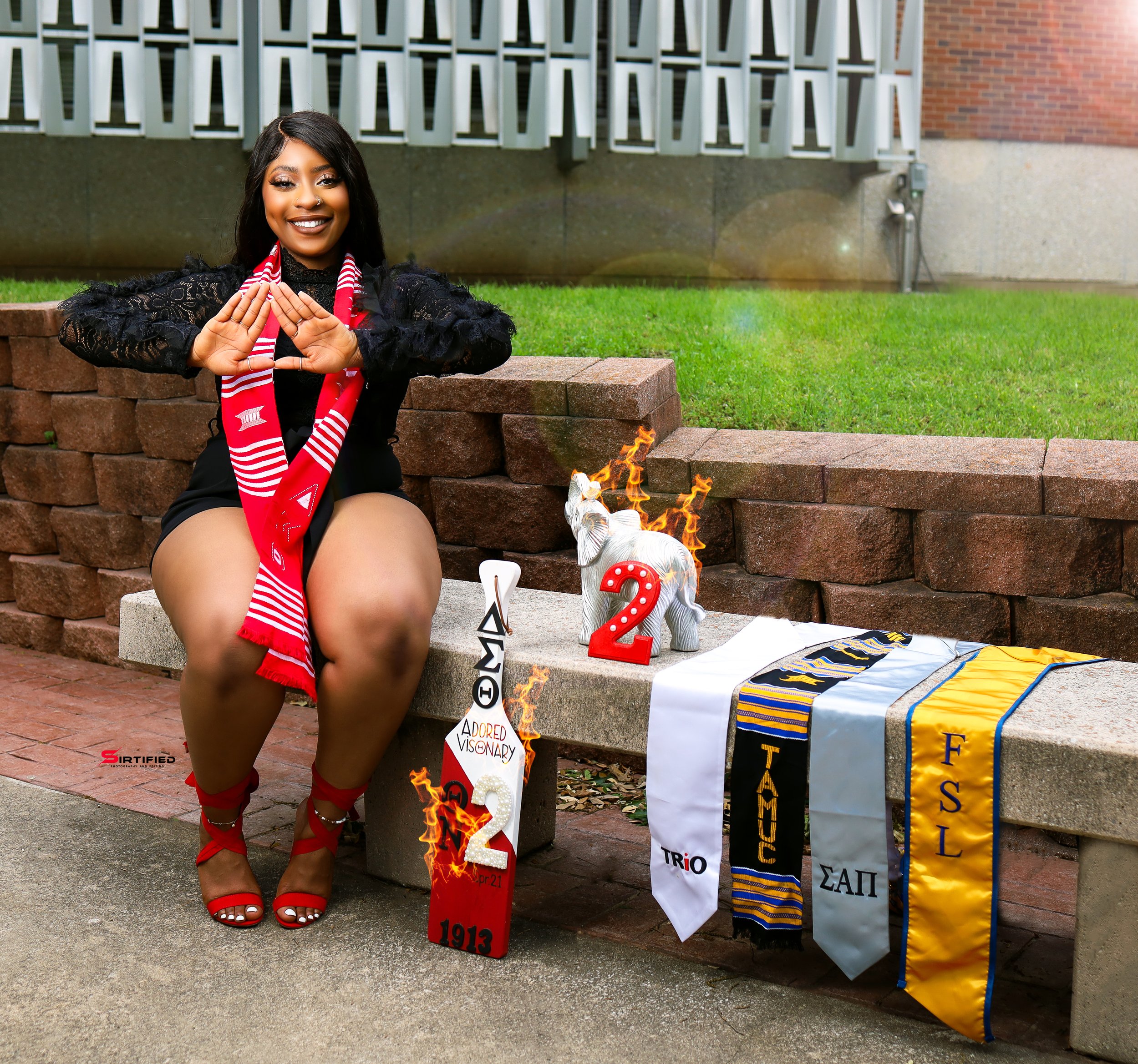 A woman sitting on a stone bench during a graduation celebration, wearing a red and white scarf, red heels, and black attire. There are decorative graduation-themed items, including a burning elephant figurine with a red number 2, and scarves from va