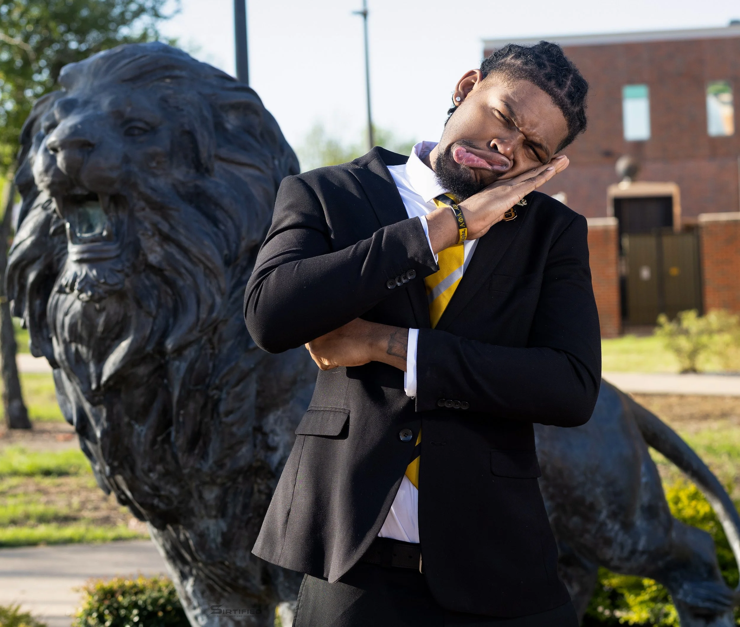 A man in a black suit and yellow tie mimics sleeping by placing his hand on his cheek and sticking out his tongue, standing outside near a lion statue.