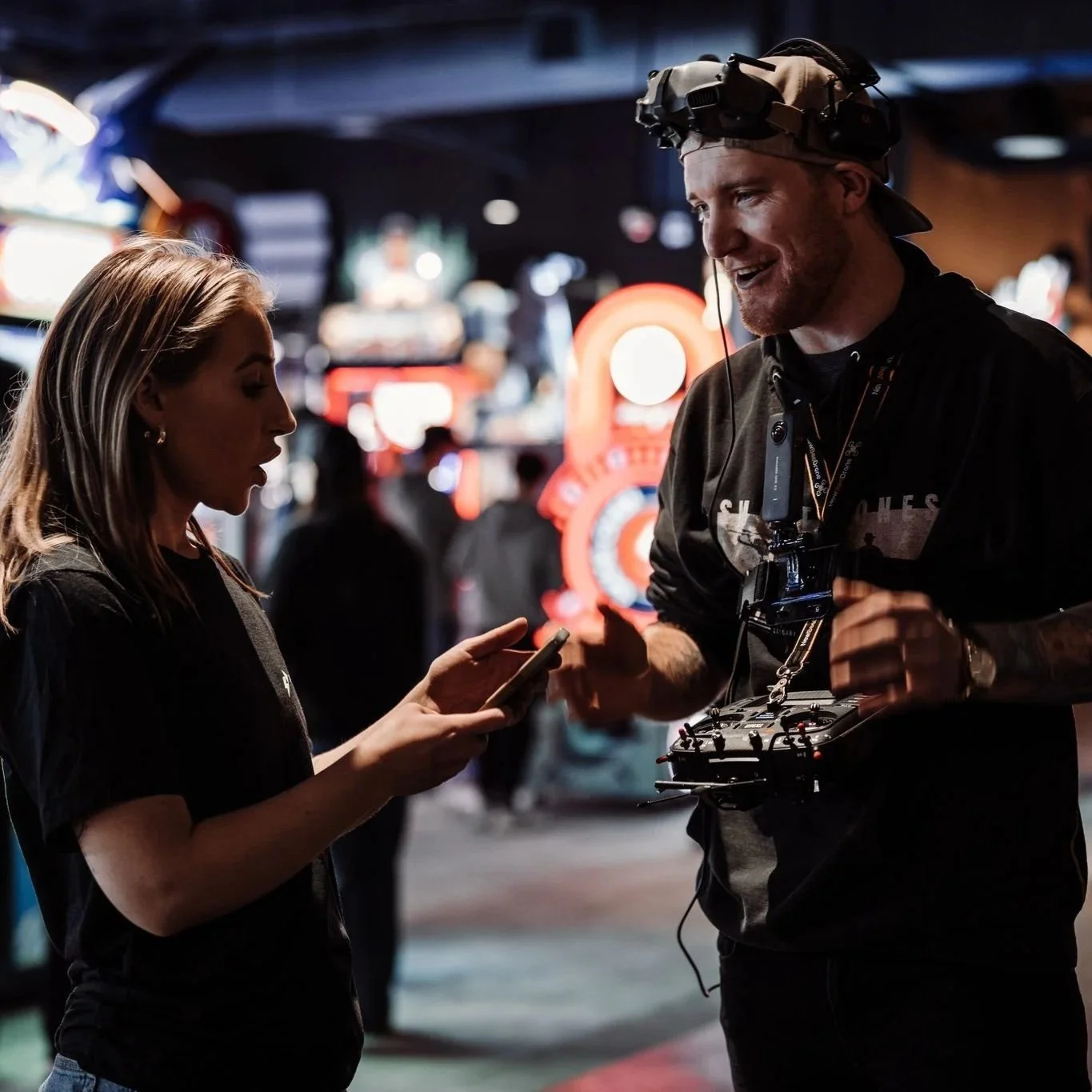 A man and woman standing in a lively, neon-lit arcade or entertainment venue. The man is wearing a headset and a drone controller around his neck, smiling and talking to the woman. The woman is looking at her phone, appearing engaged in conversation.