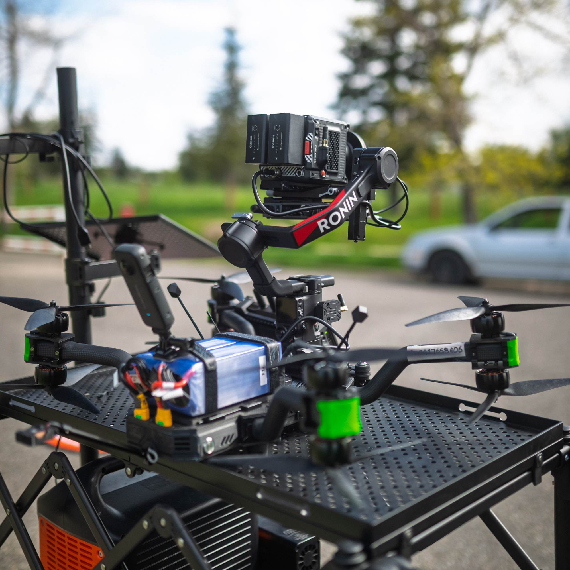 A drone equipped with a camera and various electronic components, mounted on a black platform outdoors with a blurred background of trees and parked cars.
