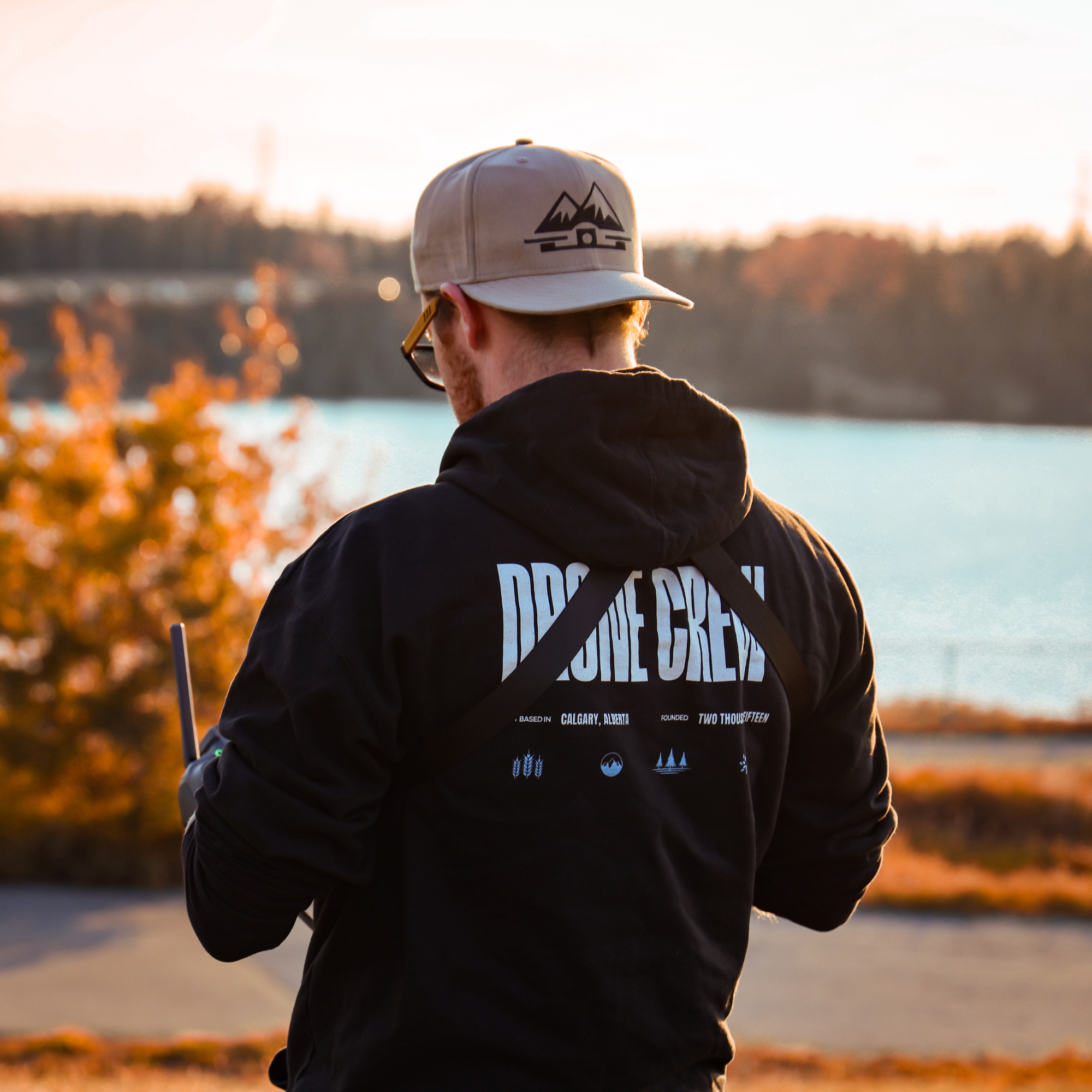 A man wearing a beige cap and a black hoodie with text on the back, standing outdoors near a body of water during sunset.